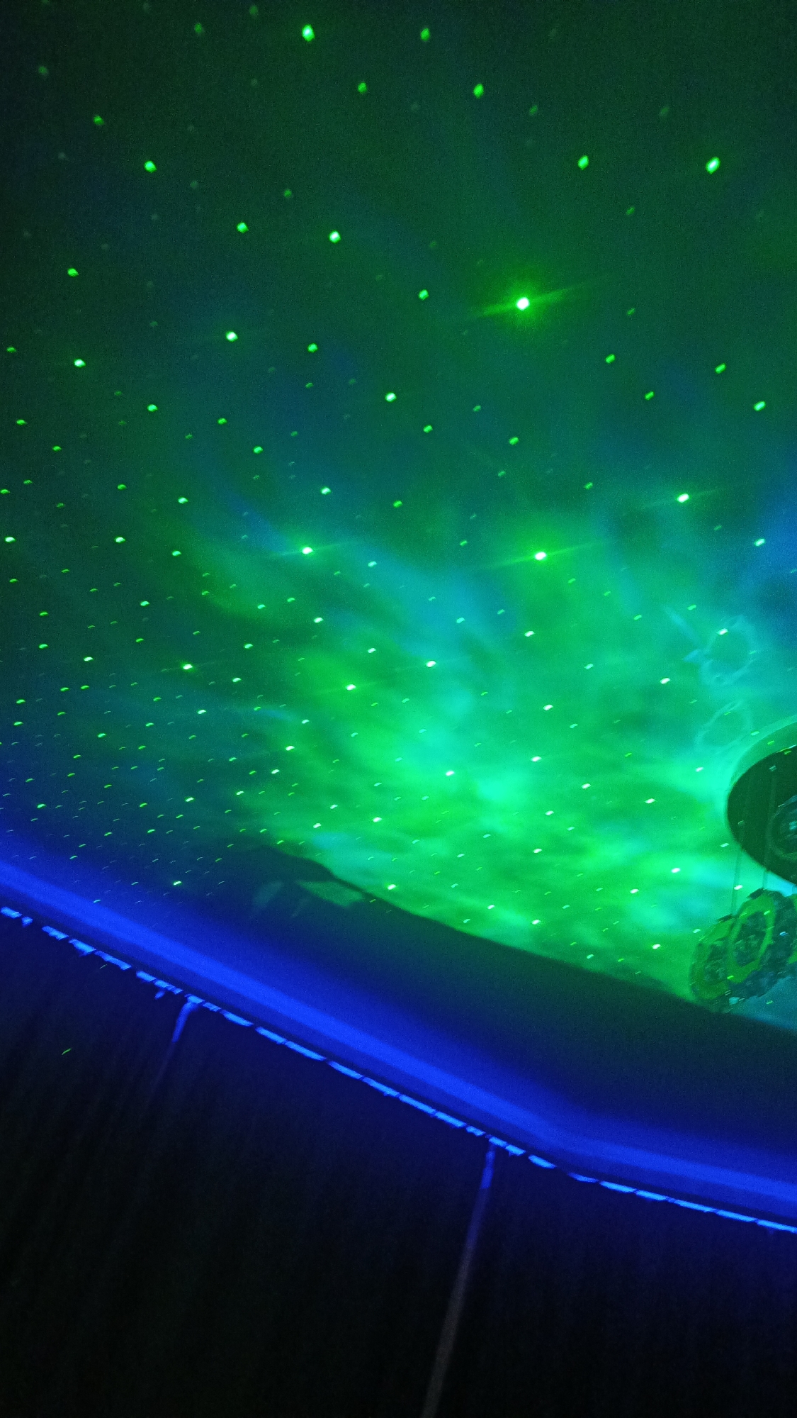 The image captures a ceiling adorned with a galaxy projection, featuring numerous small green lights against a backdrop of swirling green and blue nebulous patterns. The lower portion of the image is dominated by a dark, curved surface, likely a wall or structure, with a prominent blue light strip running along its edge. The scene appears to be indoors, possibly in a planetarium or entertainment venue. The presence of the blue light strip and the general aesthetic suggests a modern installation. The location in Zvenigorod, Russia, is not visually evident from the image itself.
