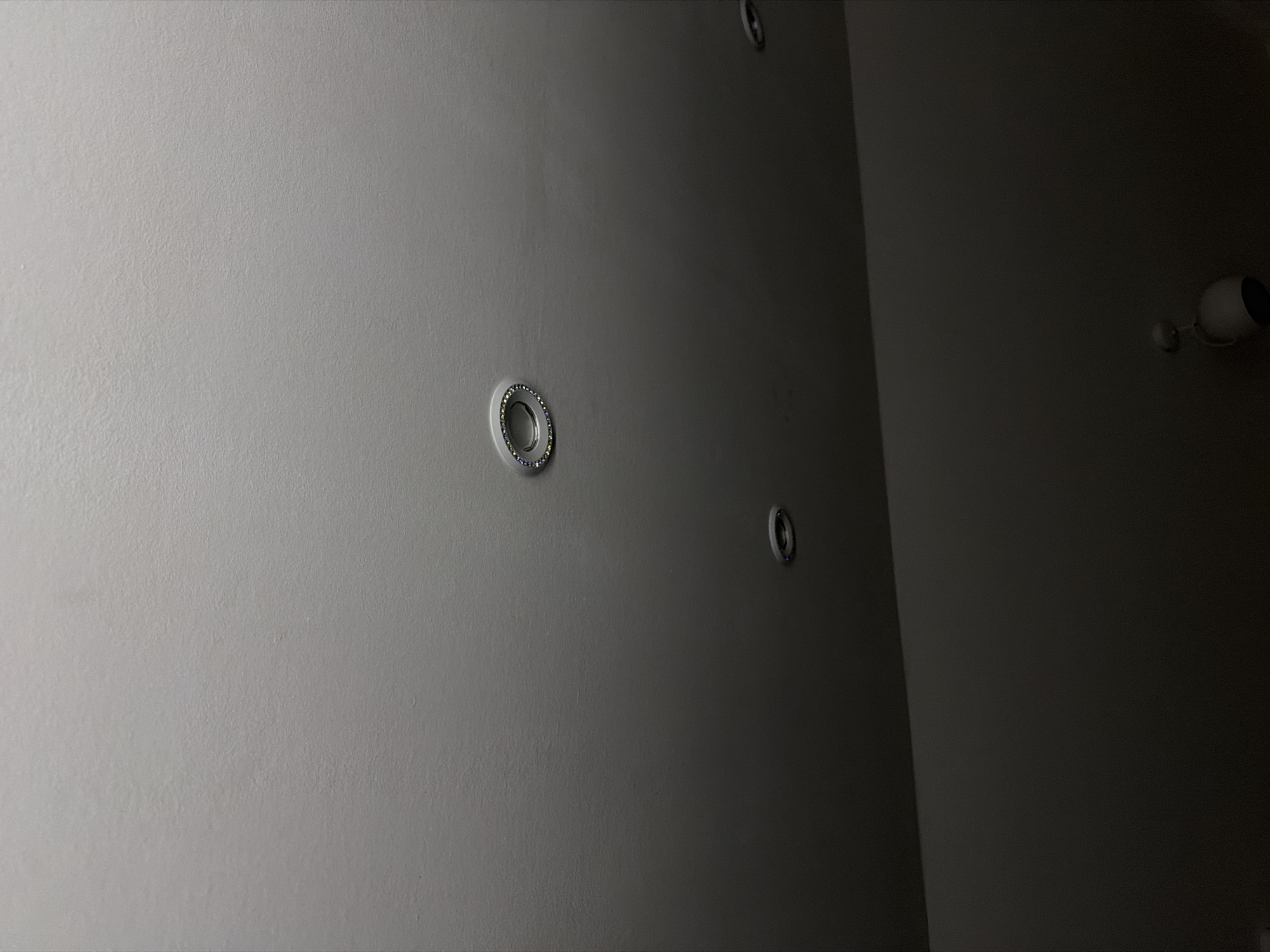 The image shows a ceiling with three recessed light fixtures. One fixture in the upper center has a decorative ring with blue and yellow embellishments. The other two fixtures are dark and less distinct. A portion of a white light fixture, possibly a ceiling fan or a pendant light, is visible in the lower right corner of the frame. The overall lighting suggests an indoor environment with a significant portion of the scene in shadow. The location is not specified in the image or the provided context.