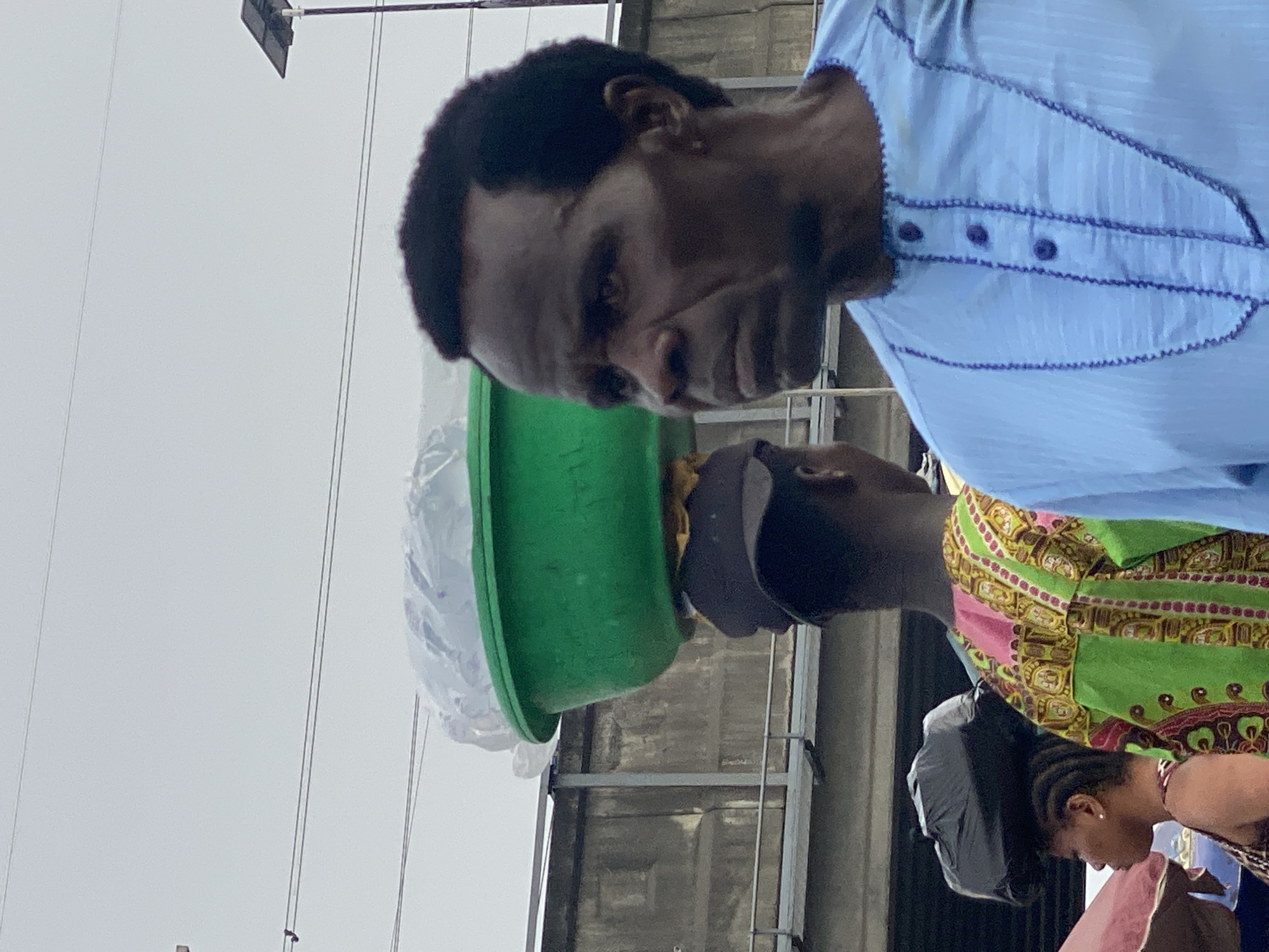 The image captures an outdoor street scene under an overcast sky, likely in Lagos, Nigeria. The primary subject is a man with dark skin and short, dark hair, positioned in the center-left. He wears a light blue traditional shirt, possibly an agbada or similar, adorned with purple embroidery along the placket and cuffs. His expression is serious or contemplative as he looks downwards. On his head, he balances a bright green plastic basin filled with items covered in clear plastic.
To the man's right, another individual is partially visible, their face largely obscured by a dark object carried on their head. This person is dressed in a vibrant, patterned garment featuring green, pink, yellow, and brown colors, typical of West African textiles. In the bottom right corner, the side profile of a third person, possibly a woman with braided hair, is visible, also looking downwards.
The background features a light grey, overcast sky traversed by several utility wires. Below these, indistinct industrial or building structures with metallic elements can be seen. A dark, broom-like object on a pole is visible in the upper left section of the frame. The scene portrays a snapshot of daily life, possibly involving street vendors or market activity. No text is visible in the image.
