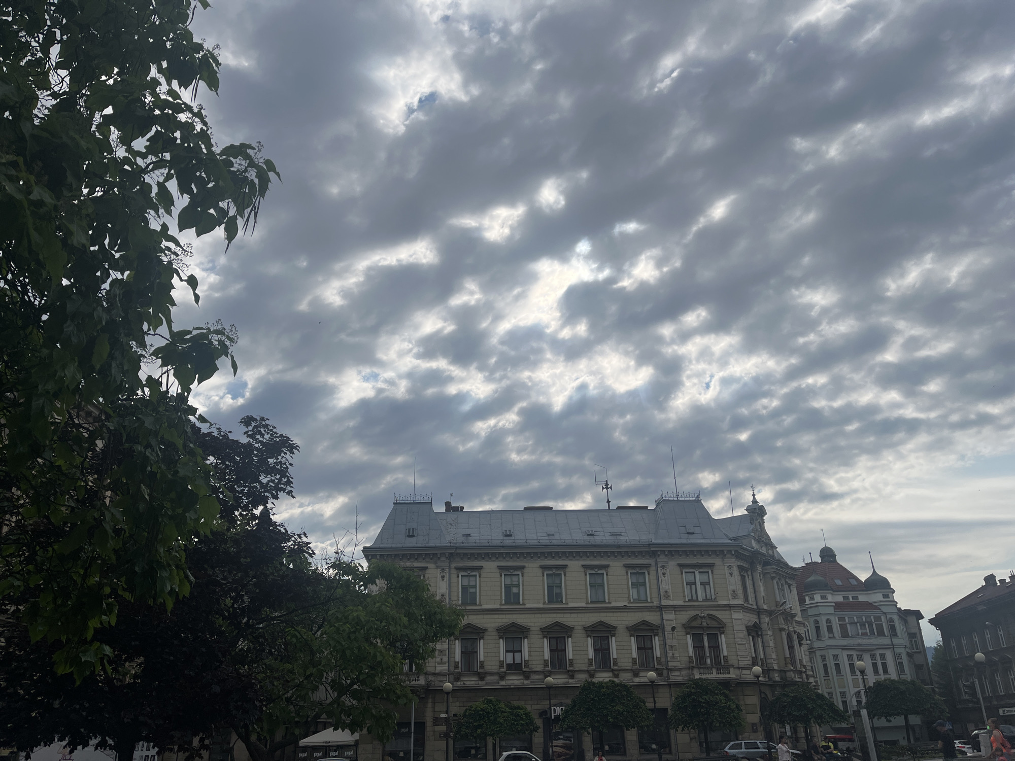 A wide shot captures a street scene in an urban environment under a cloudy sky. In the foreground, lush green trees with dark leaves partially obscure the view of buildings and the sky. The sky is filled with dramatic, textured clouds, with patches of brighter light suggesting the sun is attempting to break through.
The main focus of the image is a row of historic buildings. The largest building, located centrally, is a multi-story structure with a light-colored facade, numerous windows, and a distinctive mansard roof with dormers. A decorative pinnacle caps one of its towers. To its right, other buildings with varying architectural styles are visible, including one with a red-tiled roof and domed turrets, and further down the street, darker buildings.
The street level appears to be a public space with lampposts and manicured trees lining the sidewalk. A few cars are parked on the street. The presence of people is minimal, with only a few figures visible in the distance, suggesting a relatively calm atmosphere. The overall impression is of an old European city with grand architecture under a dynamic, overcast sky.