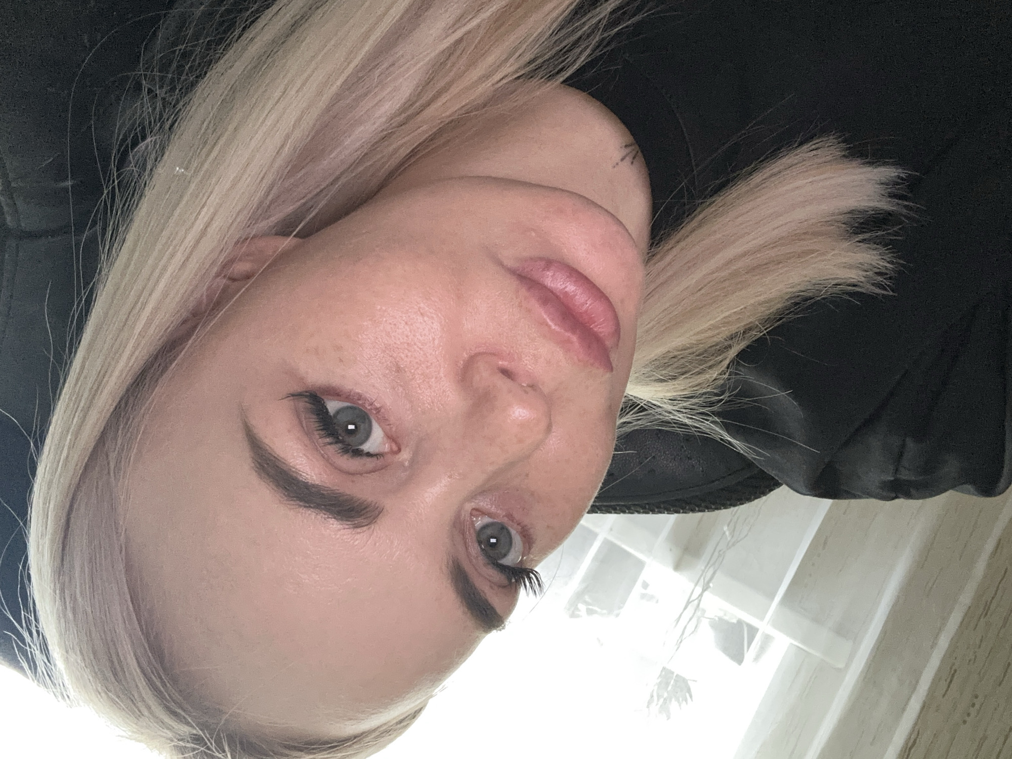 A close-up, slightly angled, candid selfie shows a fair-skinned woman with blonde hair and light blue eyes. She is wearing a black top. Her makeup includes mascara and possibly false eyelashes, and her eyebrows are well-defined. Her lips are slightly parted and appear to be wearing a light pink lipstick. The background shows a white, tiled surface and a bright light source to the left, suggesting an indoor setting near a window.