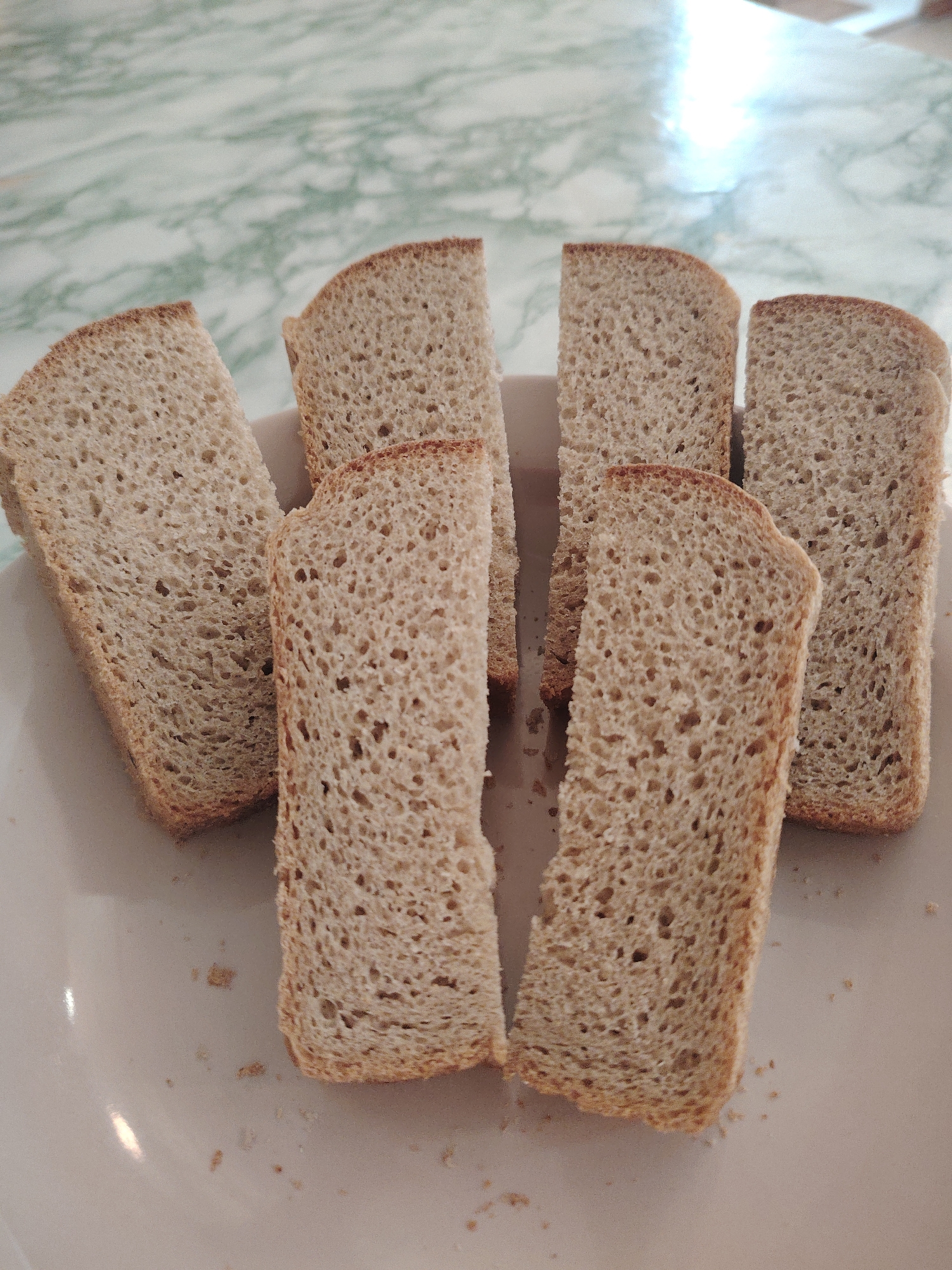 The image shows six slices of bread arranged on a white plate. The bread slices are light brown with a porous texture. Some crumbs are visible on the plate. The background is a table with a light green and white marbled surface. The location in the city of Sarapul, Russia, cannot be determined from the image.