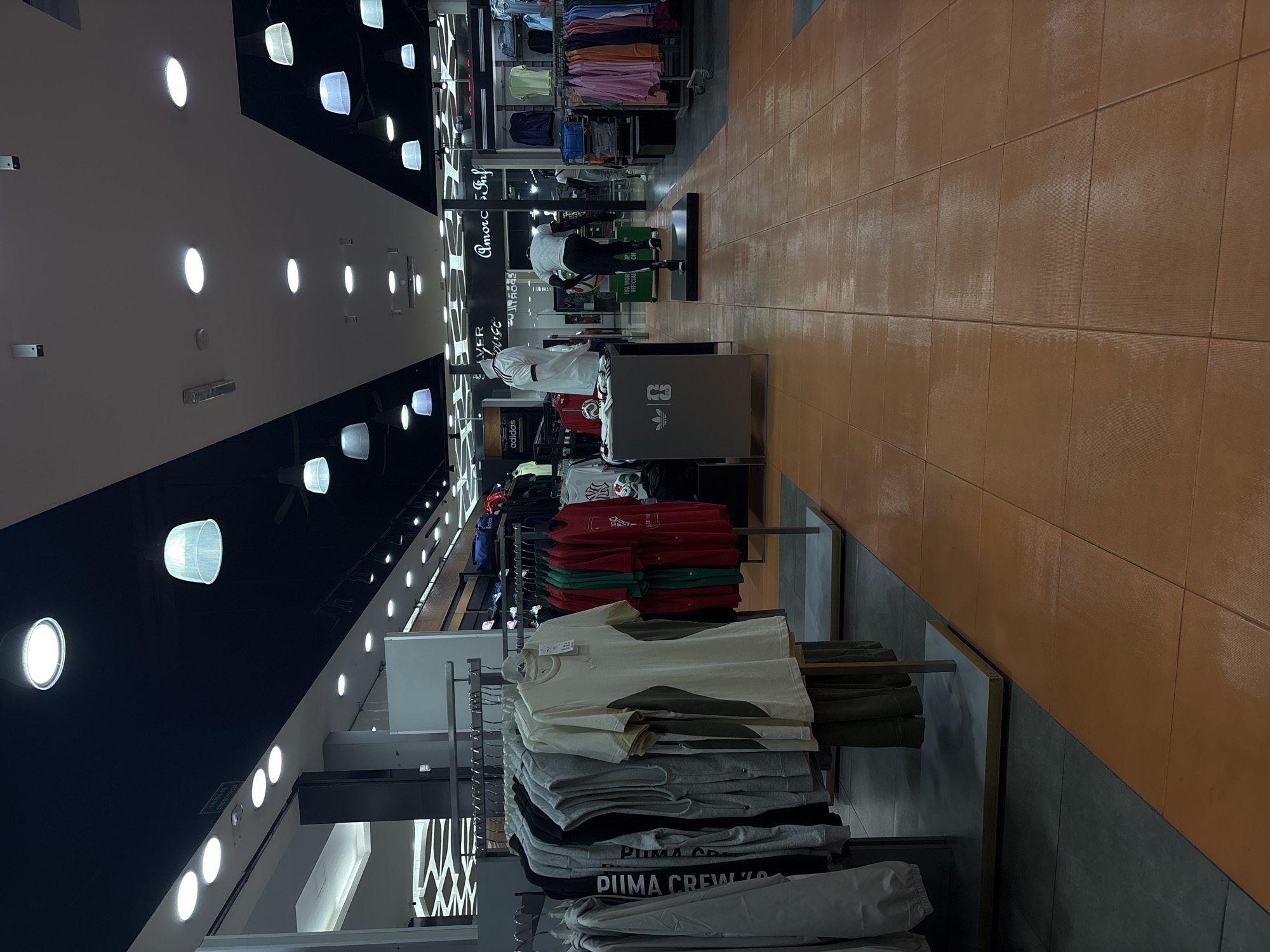 This is an interior shot of a retail clothing store, likely located in San Bernardino Tlaxcalancingo, Mexico. The store is characterized by a striking color scheme with a black and white ceiling lined with rows of circular lights, and a floor tiled in brown squares. Racks of clothing are arranged throughout the store. The garments are mostly folded and stacked on shelves. Puma and Adidas brands are visible. There are mannequins set up to display clothing as well.
