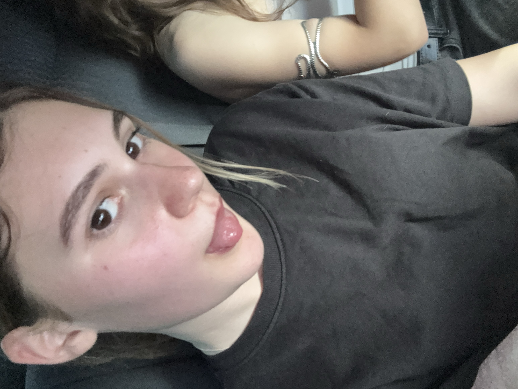 The image is a close-up selfie of a young person with light skin, brown eyes, and light brown hair. The person is wearing a dark t-shirt and has their tongue sticking out. A portion of another person with light skin and long brown hair is visible on the right side of the frame, wearing a silver arm bracelet. The background appears to be dark and textured, possibly upholstery. There is no discernible information in the image or the provided crops that would indicate a location in Koprivshtitsa, Bulgaria.