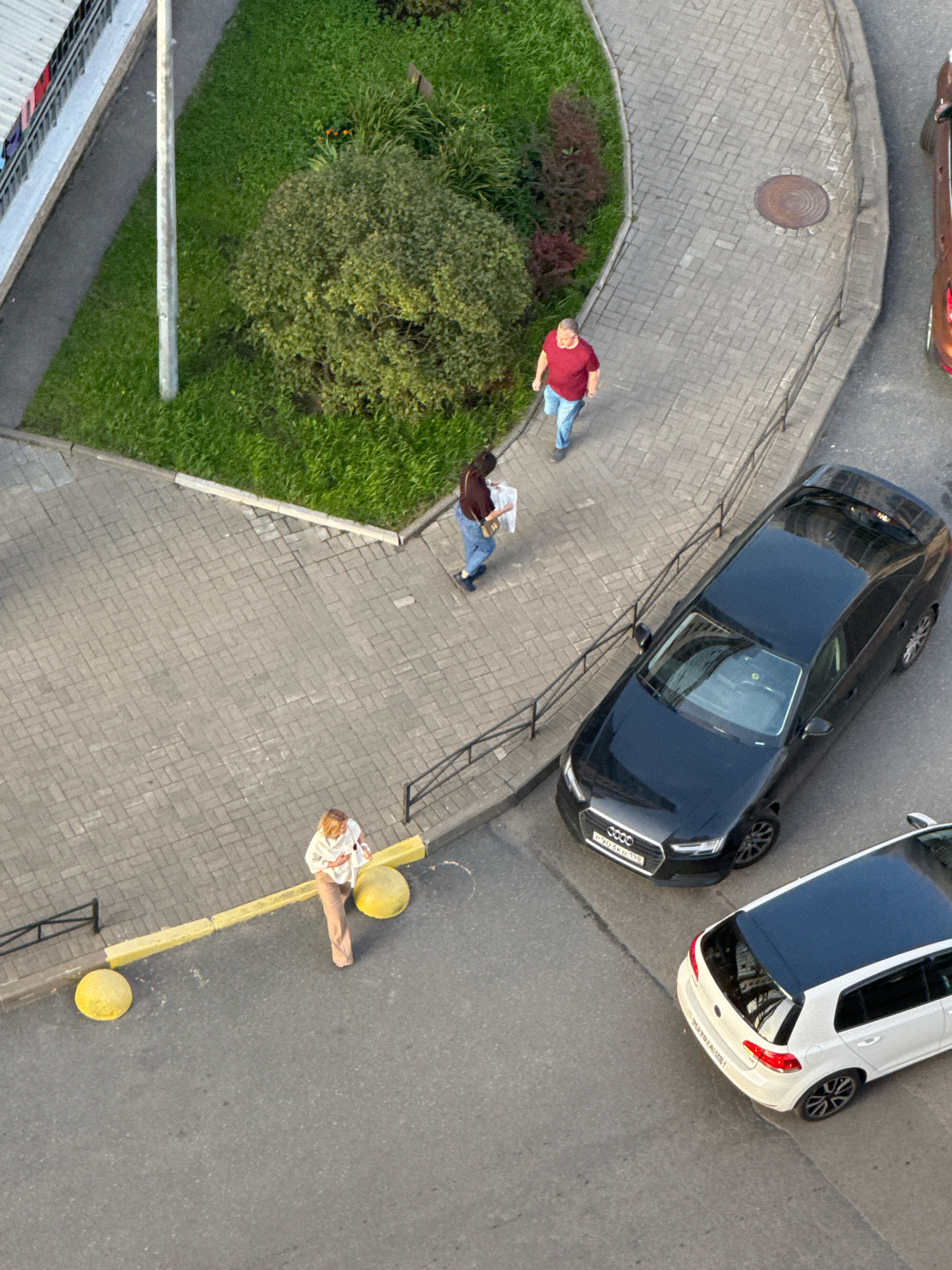An aerial shot captures an outdoor scene in an urban environment, likely Russia, with paved pathways and a grassy area. In the upper left, part of a building with a white facade and a corrugated metal roof is visible, with laundry hanging on a balcony. A concrete light post stands to the right of the building.
Several individuals are present:
- A man in a red t-shirt and blue jeans walks on the pathway.
- A woman wearing a dark top and blue jeans, carrying a white paper, walks in the same direction as the man.
- A woman in a light-colored top and beige pants stands near a yellow barrier and two round yellow bollards.
Two cars are parked on the asphalt. The first is a black Audi sedan, license plate visible. The second is a white Volkswagen hatchback with a black roof, also with a visible license plate. The pathway is made of light-colored interlocking bricks. Green grass and some shrubs are present to the left of the pathway. A manhole cover is set into the pathway.