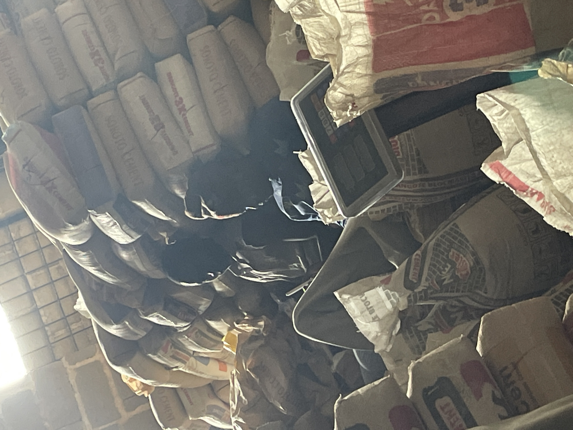 The image depicts an indoor scene, likely a warehouse, filled with stacks of cement bags. Two individuals, appearing to be young men, are situated among the bags. One individual is seated, leaning back, with their legs extended, looking down at a tablet or calculator-like device resting on a bag. The second individual is also seated, facing away from the camera, with their head bowed. The lighting suggests artificial illumination, possibly from overhead fixtures, casting shadows and highlighting the texture of the cement bags. The environment appears somewhat cluttered with the large volume of bagged materials. The presence of "SONOTO CEMENT" branding is visible on some of the bags, indicating the type of product stored. The overall impression is one of a storage facility where inventory management or a brief respite is occurring. The prompt specifies the location as the city of Jos, Nigeria.