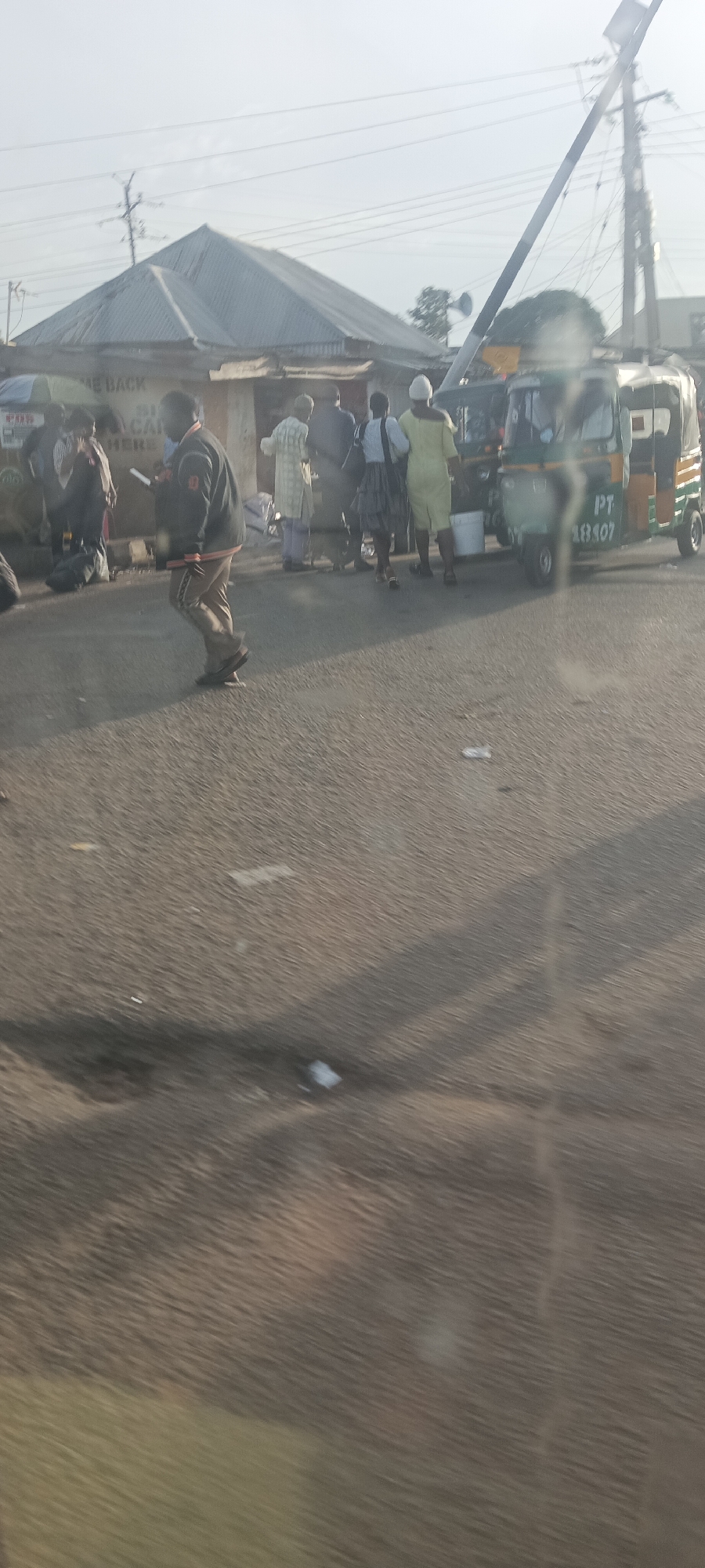 This image captures a vibrant roadside scene in Gura Top, Nigeria, likely during the early morning or late afternoon given the soft, slightly hazy sunlight and visible shadows on the asphalt road.
The main subjects are several individuals congregated near a building and two tricycles, commonly known as Keke NAPEP in Nigeria. In the foreground, a person wearing a dark jacket and light-colored trousers is captured mid-stride, moving from left to right across the road. Other people are standing or walking near the roadside building and the tricycles. One person in a light yellow garment stands near a tricycle. Most individuals appear to be engaged in routine activities, possibly waiting for transport or interacting at a local business.
The setting includes a one-story building with a corrugated metal roof, suggesting a commercial establishment. Partial text is visible on its facade, including "ME BACK" and "SI CAB," possibly part of a sign like "WELCOME BACK" or "STATION CAB." Overhead, power lines and utility poles crisscross the sky, typical of an urban or semi-urban area.
A prominent object is a green and yellow tricycle parked on the right, displaying the text "PT 18407" on its side. Another tricycle is partially visible behind it. A white bucket is placed on the ground near the tricycles. The road surface is asphalt, with some small pieces of litter scattered on it. The overall atmosphere is one of daily life unfolding at a local hub.