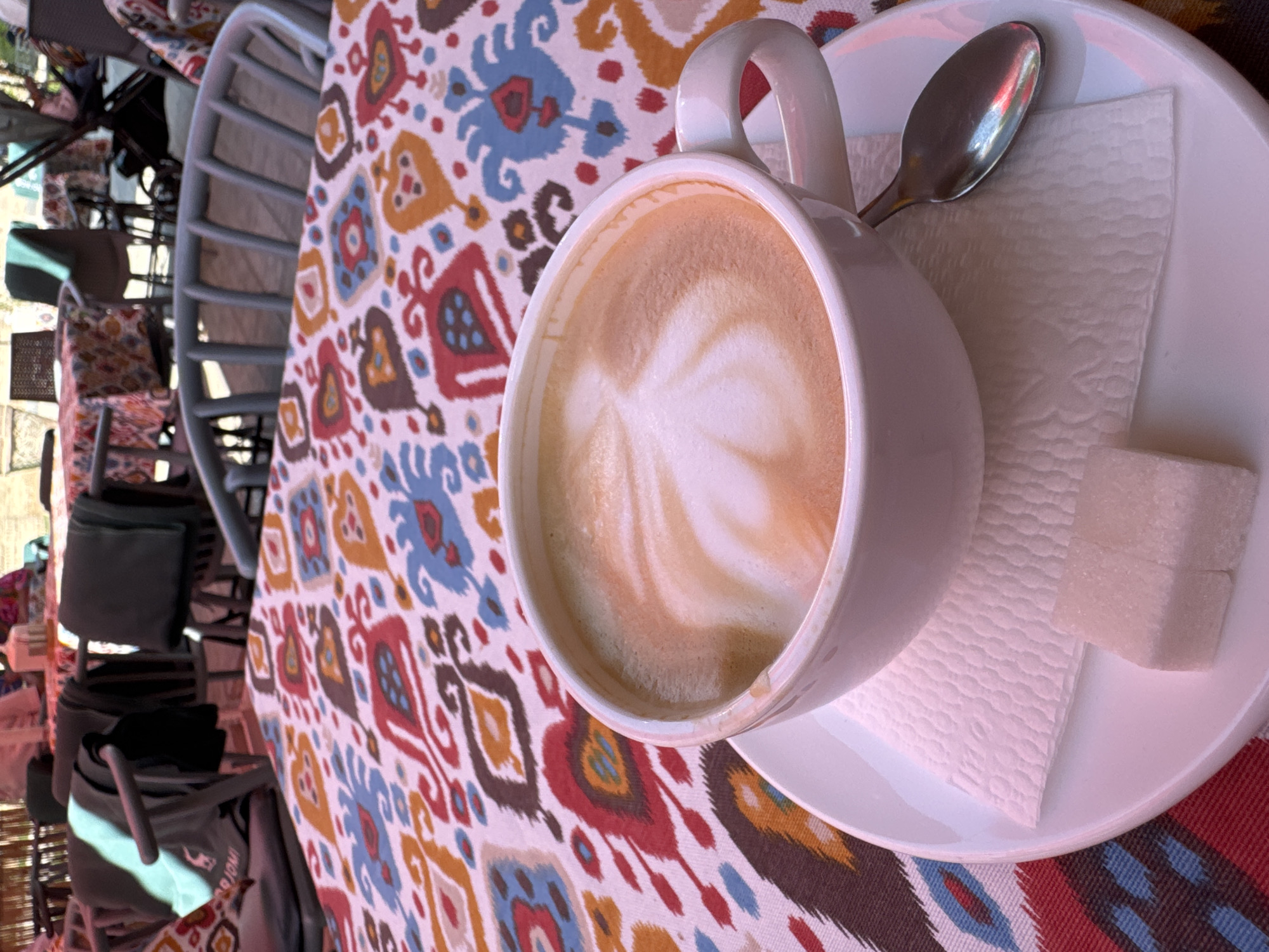 A latte sits on a white saucer with a spoon and a sugar cube, on a table covered with a colorful ikat fabric. The table is surrounded by grey metal chairs. The setting appears to be an outdoor cafe in Samarkand City, Uzbekistan, indicated by the ikat fabric which is characteristic of the region. Other tables and chairs, possibly other cafe patrons, can be seen blurred in the background.