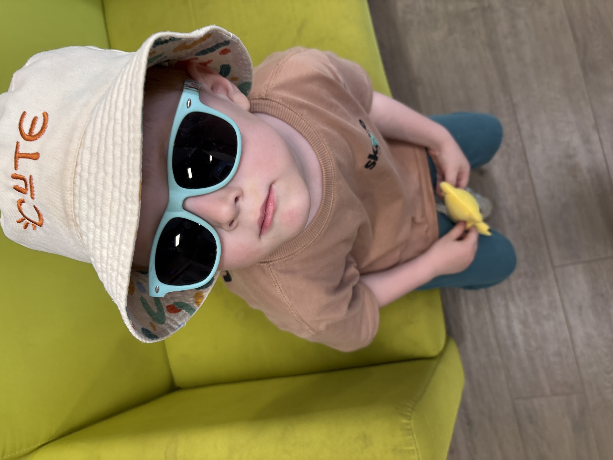A fair-skinned child wearing a light tan T-shirt with a small graphic, teal pants, a white bucket hat with "cute" embroidered on it, and light blue sunglasses sits on a bright green sofa. The child is holding a small yellow object. The location is indoors with a light wood-colored floor. The location in the city of Izhevsk, Russia cannot be determined from the image.