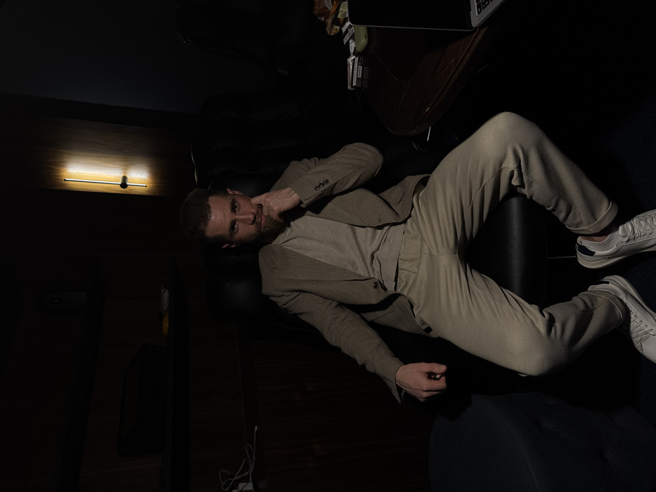 A man reclines on a black leather sofa, wearing a beige suit, t-shirt, and white sneakers. He rests a finger near his temple, gazing toward the viewer. A dark wooden table with items sits nearby. A linear light fixture illuminates a section of the dark wall. The image is dominated by dark shadows.