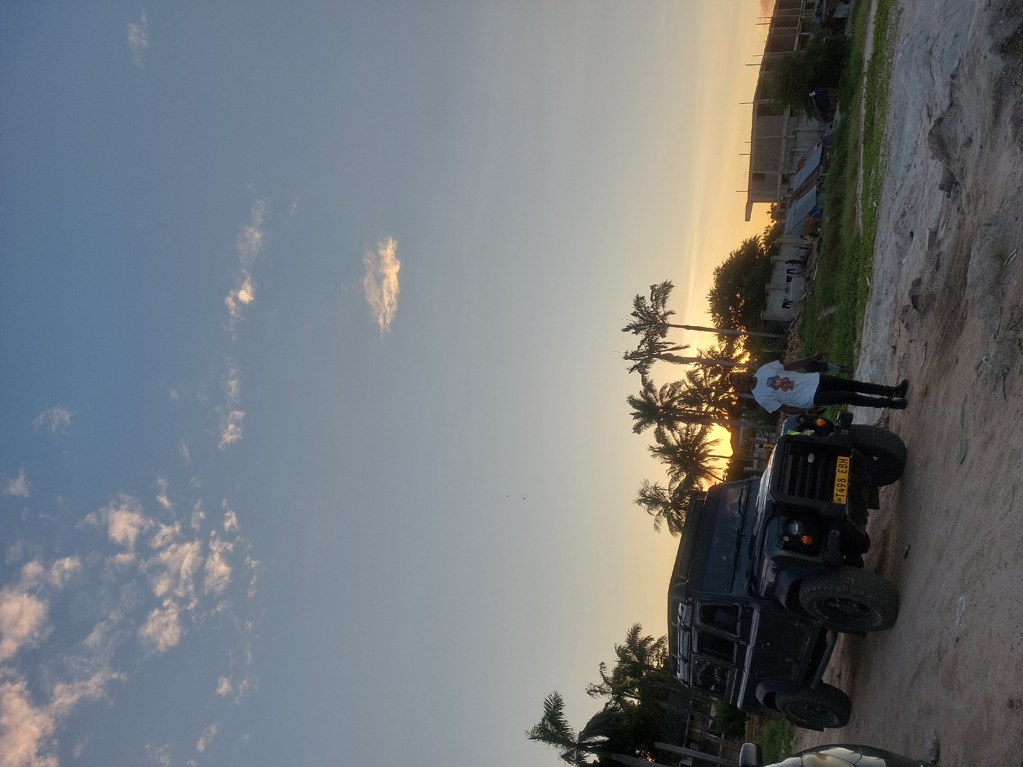 A man stands beside a black Land Rover Defender 110. The vehicle has a Tanzanian license plate, "T 498 EBH". Palm trees are behind them. The sky is a gradient of blue to orange, indicative of either sunrise or sunset. In the background, there's a partially constructed building. The ground is sandy. The photo appears to be taken in Dar es-Salaam, Tanzania, given the license plate.