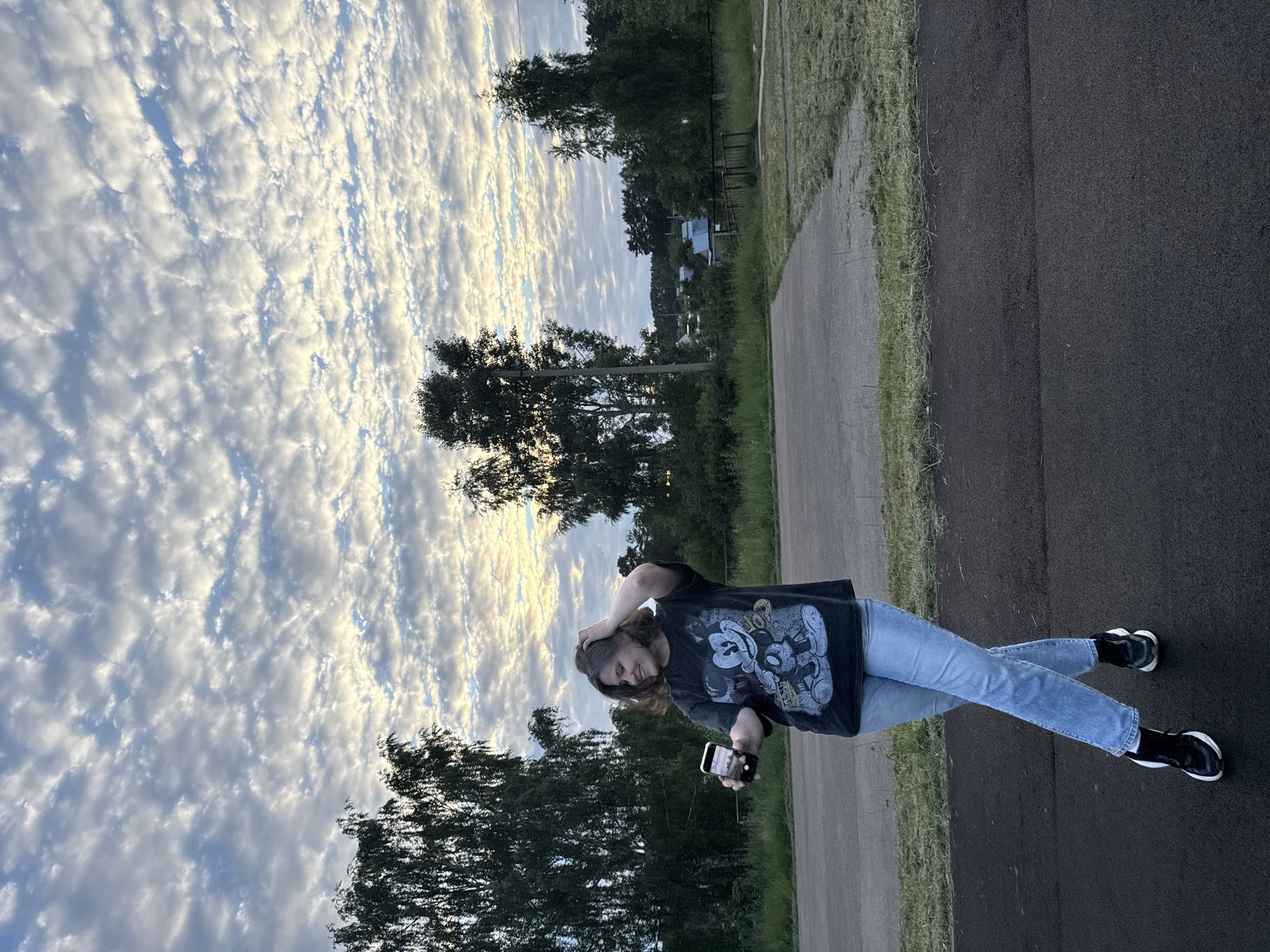 A young woman with short brown hair stands outdoors, taking a selfie with her smartphone. She is wearing a black t-shirt with a graphic print of Mickey Mouse and blue jeans. Her left hand is raised to her head, and her right hand holds the phone. She is positioned on a paved area, with a grassy field and trees in the background. The sky above is filled with a dramatic display of white and grey clouds, with patches of blue visible. The scene appears to be located in a semi-rural area, possibly on the outskirts of Chaikovsky, Russia, given the presence of trees and what appear to be some rural dwellings in the distance. The lighting suggests it is either early morning or late evening.