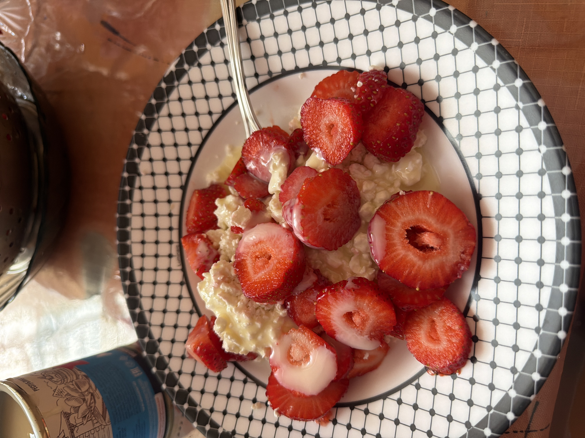 A plate with a black and white patterned rim is filled with sliced strawberries and cottage cheese. A metal spoon rests on the plate. The plate sits on a table with a textured, brown surface. A can with a blue label is partially visible in the lower-left corner. The scene appears to be indoors, under natural lighting. There's no indication of the location being Moscow, Russia. There are no people visible in the image.