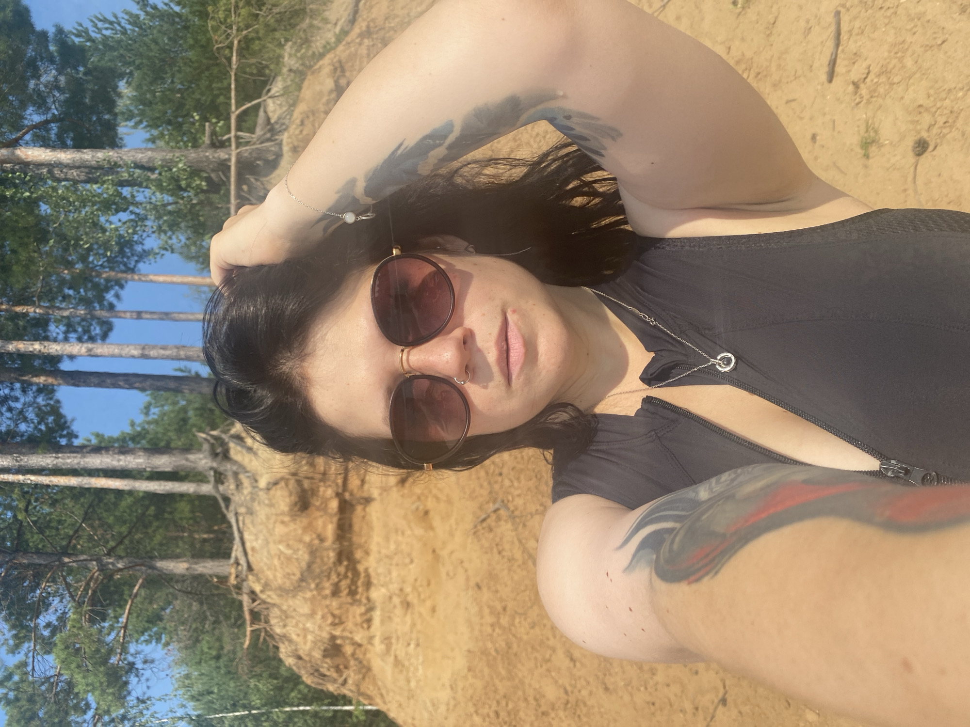 A medium shot selfie shows a woman with dark hair and tattoos on her arms, wearing sunglasses and a black zippered top. She is standing on a sandy area with a cliff face behind her. Trees are visible on top of the cliff. The sky is blue. The location is identified as Novyi, Russia.