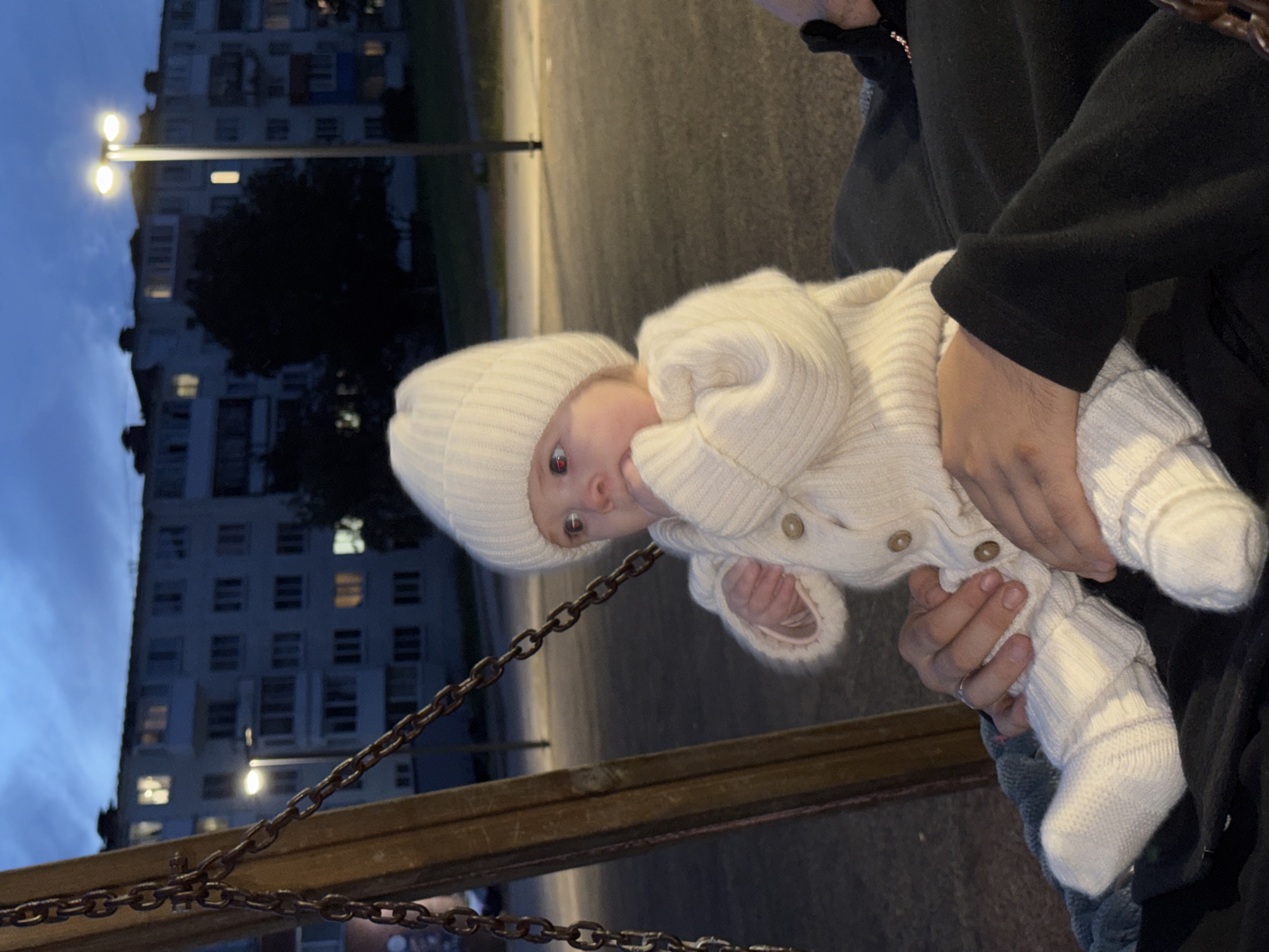 A baby wearing a white knitted hat, sweater, and socks is sitting on someone's lap, who is sitting on a swing. The baby is looking to the left and has their hand near their mouth. The background shows an apartment building, a street light, and a tree, all illuminated by the twilight sky. The image appears to have been taken in Noviy, Russia.
