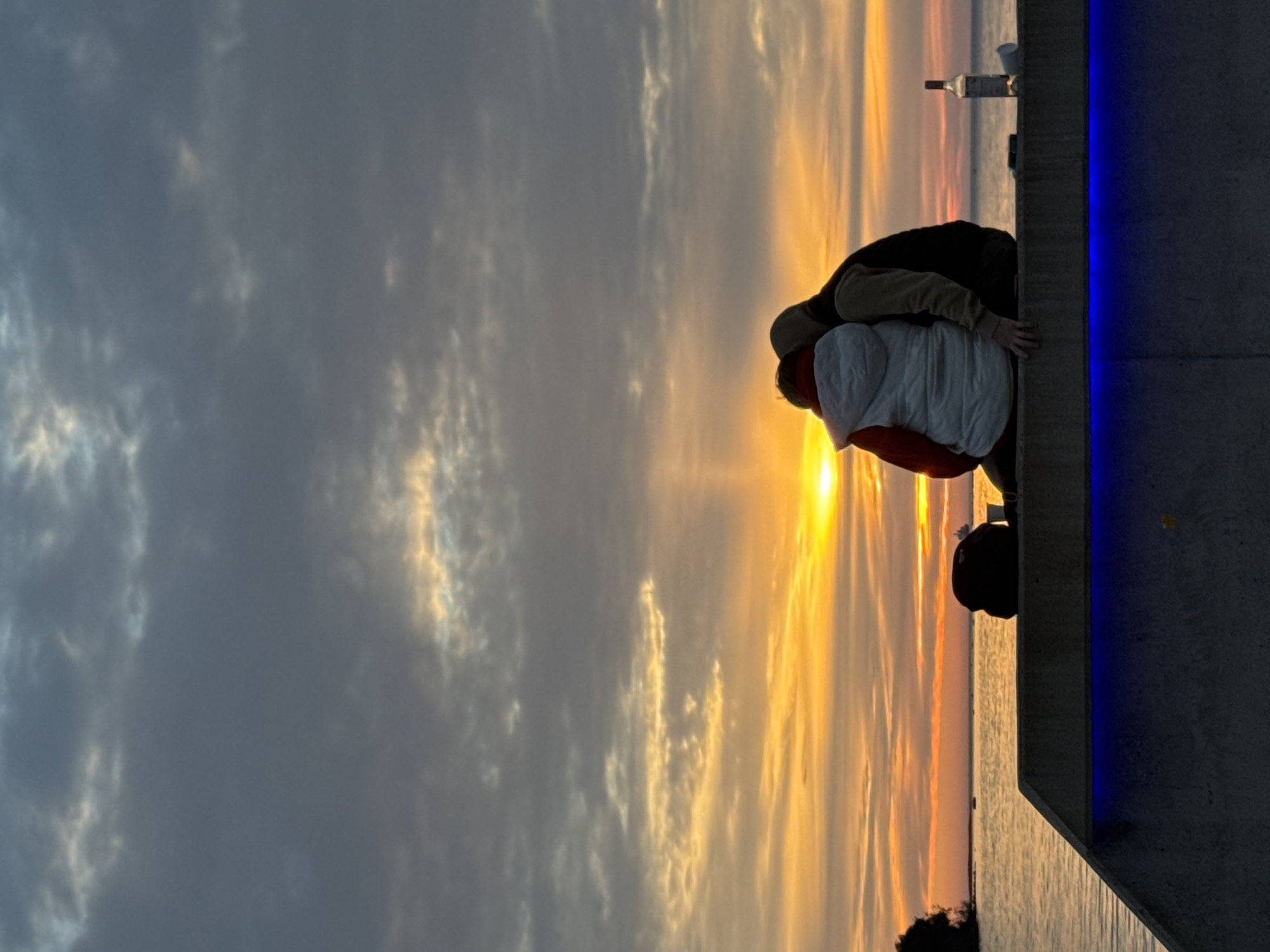 The image captures a silhouetted couple embracing on a concrete ledge overlooking the sea at sunset. The sky is filled with dramatic clouds and streaks of orange and yellow light. A bottle of wine and a cup sit on the ledge. The scene is dimly lit, with the concrete structure below illuminated by blue lights. The specific location is the pier area in Tallinn, Estonia, known for its scenic sea views and popular gathering spots for locals and tourists alike, especially during sunset.