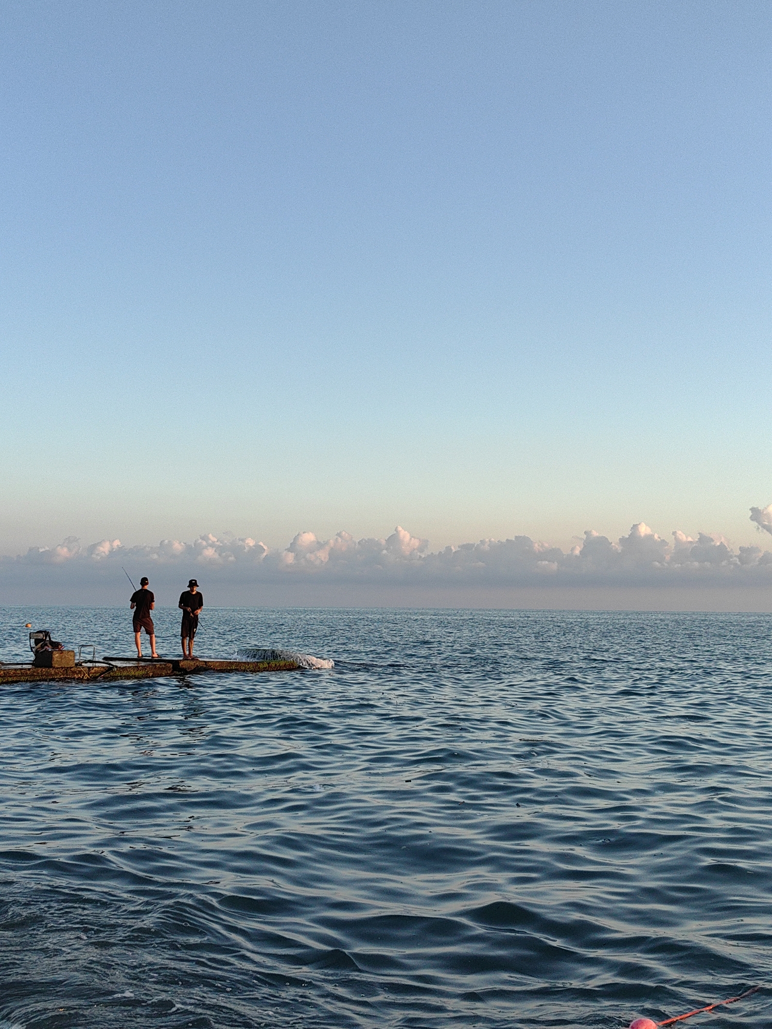 The media file captures a serene morning scene at the sea in Sochi, Russia. Two individuals, silhouetted against the soft dawn light, are engaged in fishing from a small, weathered concrete structure extending into the calm blue water. The sky above is a gradient of pale blue, transitioning from a lighter hue near the horizon to a deeper shade overhead. A line of fluffy, pink-tinged clouds stretches across the horizon, hinting at the sunrise. The sea is gently rippling, reflecting the soft colors of the sky. The overall atmosphere is one of tranquility and peaceful solitude.