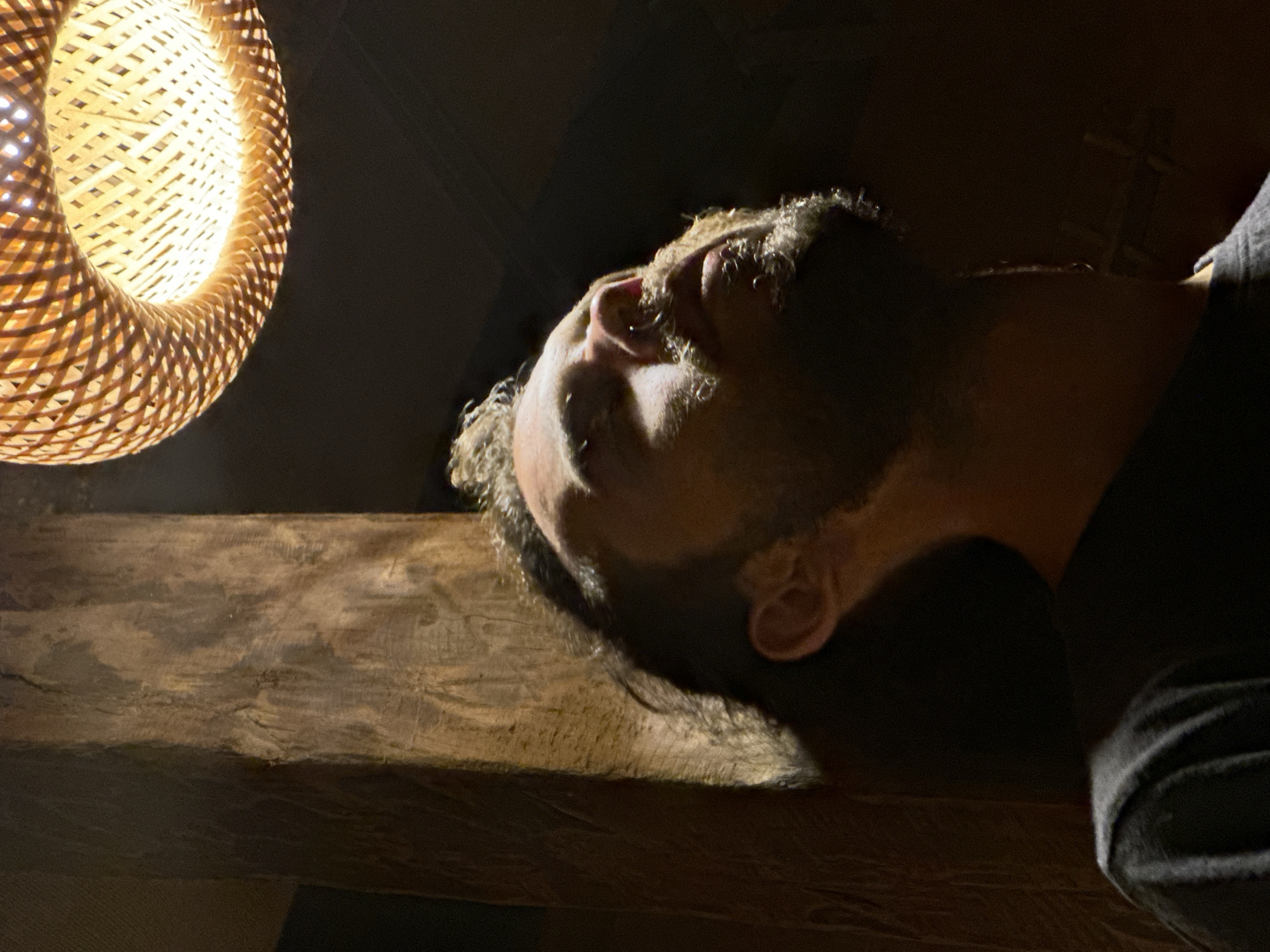 The media file presents an intimate, low-light portrait from a low-angle perspective. The primary subject is a person, likely male, seen in profile from the chest up, looking upwards. His face, particularly his nose, chin, and parts of his cheek and ear, is illuminated by a strong, warm light source, highlighting his short curly hair and stubble. The rest of his body is obscured by deep shadow, though a dark shirt is visible.
The scene features a prominent woven straw or bamboo light fixture, emitting a bright, warm yellow light. This fixture is positioned in the upper left of the frame, casting dramatic shadows and creating the primary illumination for the image. Below the person's head and extending horizontally across the lower middle of the frame is a rugged, dark wooden beam or plank, showcasing its natural grain and texture. The background and foreground are enveloped in deep shadows, suggesting an indoor setting at night or in a dimly lit room.
The person appears to be either lying down or leaning back, gazing intently towards the light source or an unseen point beyond it. The expression is contemplative or serene, with no strong emotions overtly displayed. The strong contrast between light and shadow creates a moody and atmospheric tone. There is no visible text in the image. The rustic elements like the woven lamp and wooden beam align with the suggested location context of Du Già Commune, Vietnam.