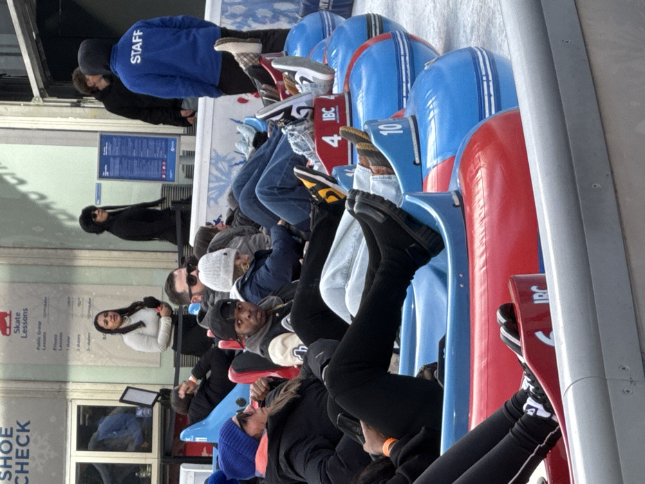 A line of people sits in individual blue and red sled-like seats. Most of the people are facing forward with their legs extended into the seats. Shoes are visible, resting on the sleds. A person in a blue "Staff" jacket stands at the front of the line. Other people are standing or walking along a wall with posted signs, including one that says "Shoe Check".