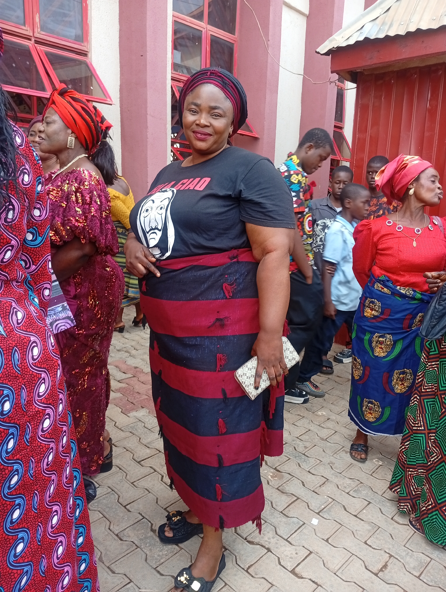In Narayi, Nigeria, a medium shot captures a group of people outside a building with red and pink facade. The central figure is a woman wearing a black T-shirt with a white mask graphic, a black and red striped skirt, and black sandals. She also wears a head wrap with red and black colors. She holds a small, light-colored clutch in her left hand. The woman stands on a paved walkway. Other people, including children and women in traditional clothing, are scattered around and behind her. The image captures a slice of everyday life in Narayi.