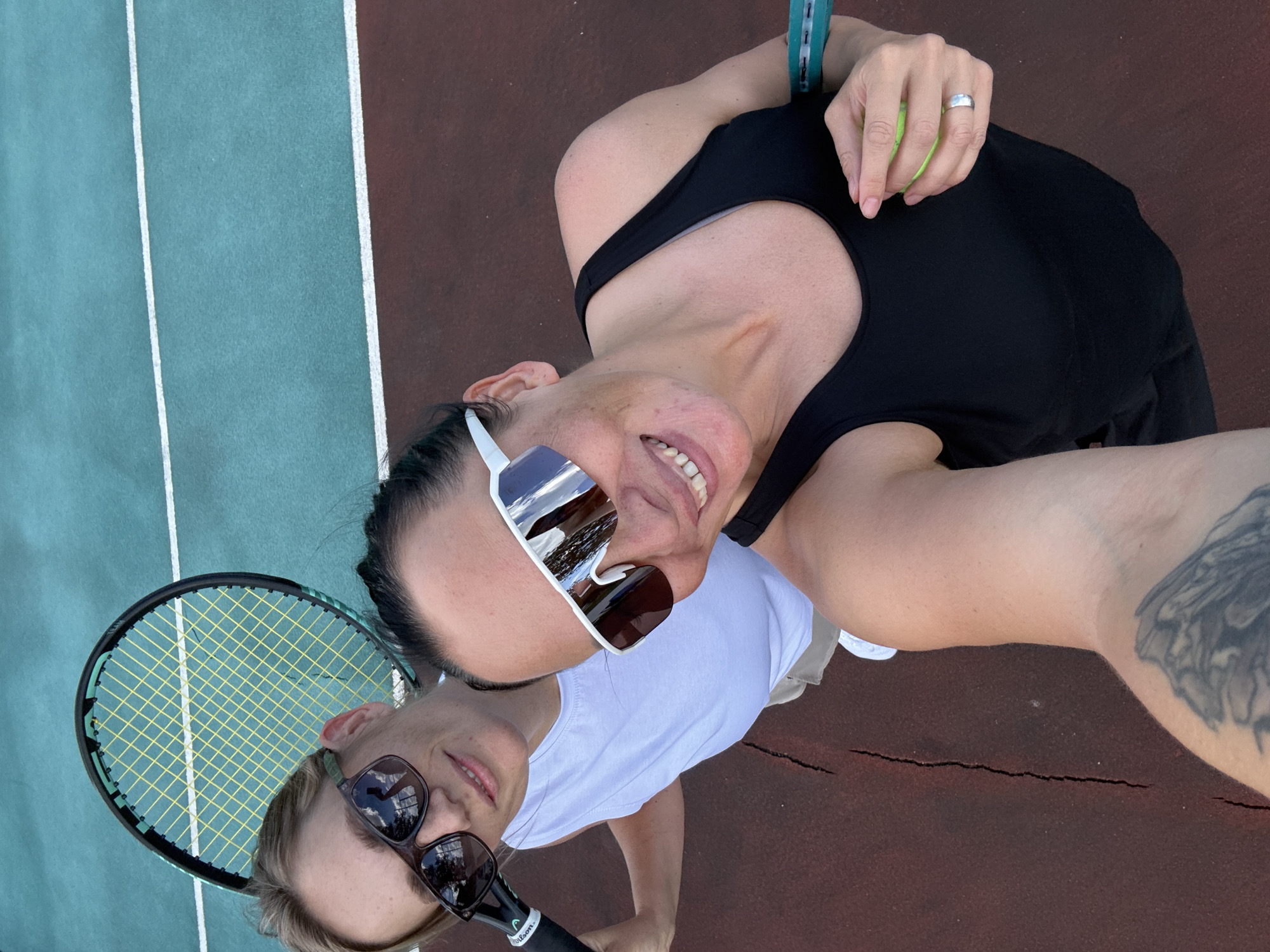 The image depicts two women on a tennis court. The woman in the foreground on the right is wearing a black tank top and large white-framed sunglasses, holding a tennis ball in her right hand. The woman on the left is wearing a white tank top and dark-framed sunglasses, holding a tennis racket. Both women are smiling and appear to be taking a selfie. The tennis court surface is a reddish-brown color with turquoise sections and white lines. The background shows the turquoise court extending upwards. The overall scene suggests a recreational activity taking place outdoors. There is no discernible information in the image that would allow for its location in Chaikovsky, Russia.