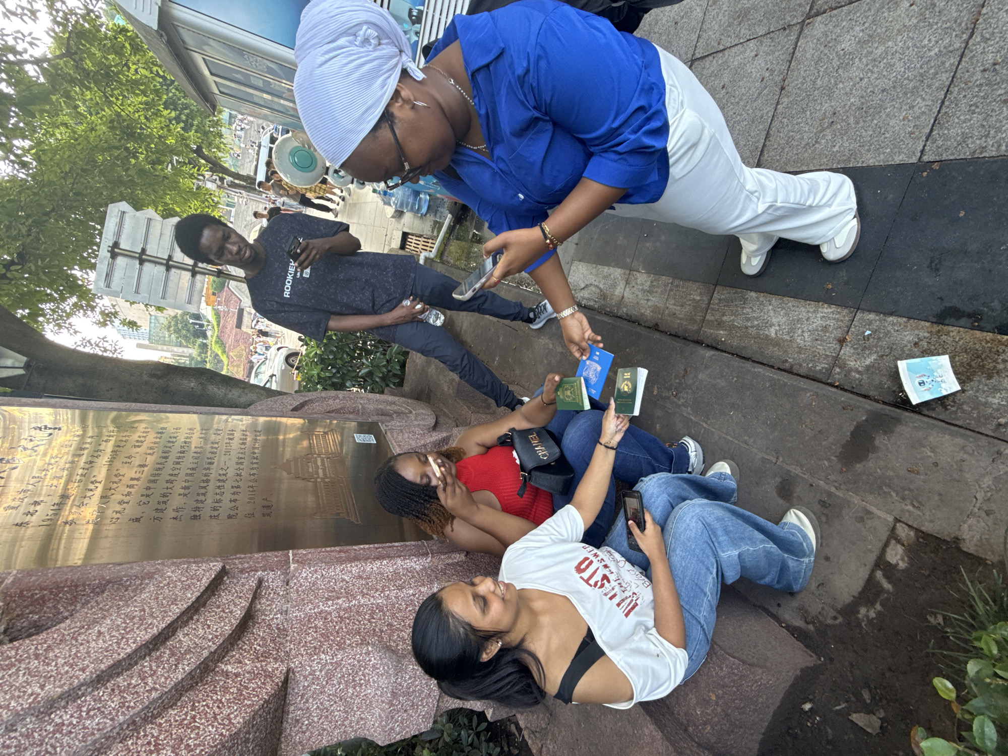 Four individuals are present in an outdoor urban environment. Two women are seated on a stone ledge, one wearing a white off-the-shoulder top and blue jeans, and the other a red tank top and blue jeans. The first woman is holding a smartphone in her left hand and looking to her right. The second woman is holding a small item to her mouth. Between the seated women are three passports, two green and one blue, on the ground. To the right of the seated women, a third individual, a woman wearing a blue long-sleeved shirt and white pants, is standing and holding a smartphone in her left hand, interacting with the passports. To the left and slightly behind the seated women, a fourth individual, a young man wearing a dark t-shirt and dark pants, is standing and looking down at a smartphone he holds in his right hand. In the background, trees, buildings, street signs, and a public transportation structure are visible. The ground appears to be a paved walkway with some dampness and debris. The stone ledge the women are seated on is part of a monument with inscribed text and an embossed architectural design, likely located in Yuzhong District, China, given the presence of Chinese characters on the monument.