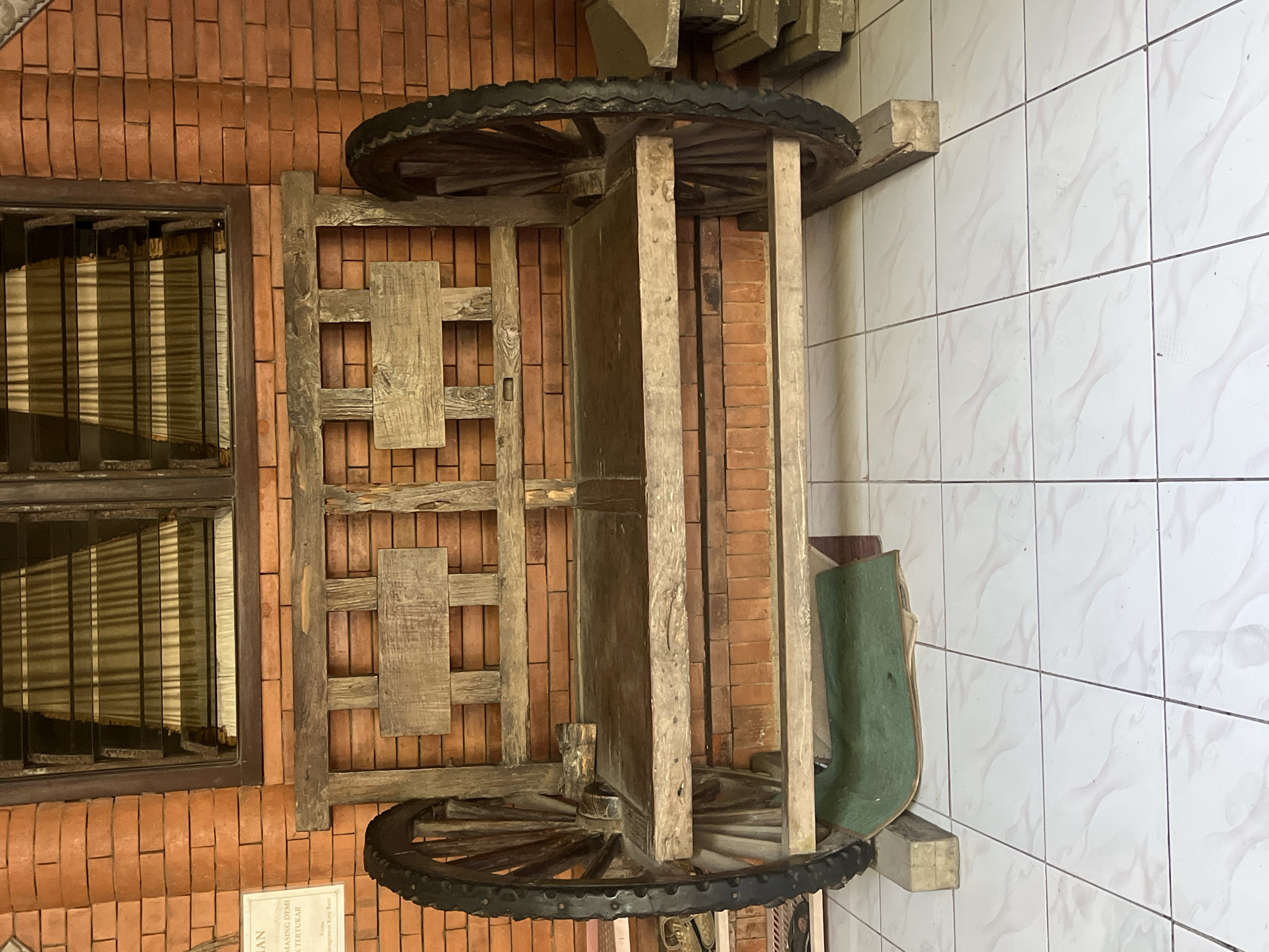 The image shows a rustic wooden bench with two large wagon wheels on either side, placed on a tiled floor in front of a brick wall and a window with horizontal louvers. A green mat is partially visible beneath the bench. A sign is affixed to the brick wall to the left of the bench. The overall setting appears to be an outdoor or semi-outdoor space, possibly a courtyard or a porch, given the tiled flooring and brickwork. The bench itself is made of rough-hewn wood, with a slatted backrest and seat, and two wooden panels on the back. The wagon wheels appear to be old and weathered, with rubber tires. The window above the bench has curtains visible through the louvers. The scene is static and devoid of human presence. Without further context, the specific location within Jepara, Indonesia, cannot be determined, though the architectural style might suggest a traditional or artisanal setting often found in such areas known for their crafts and cultural heritage.