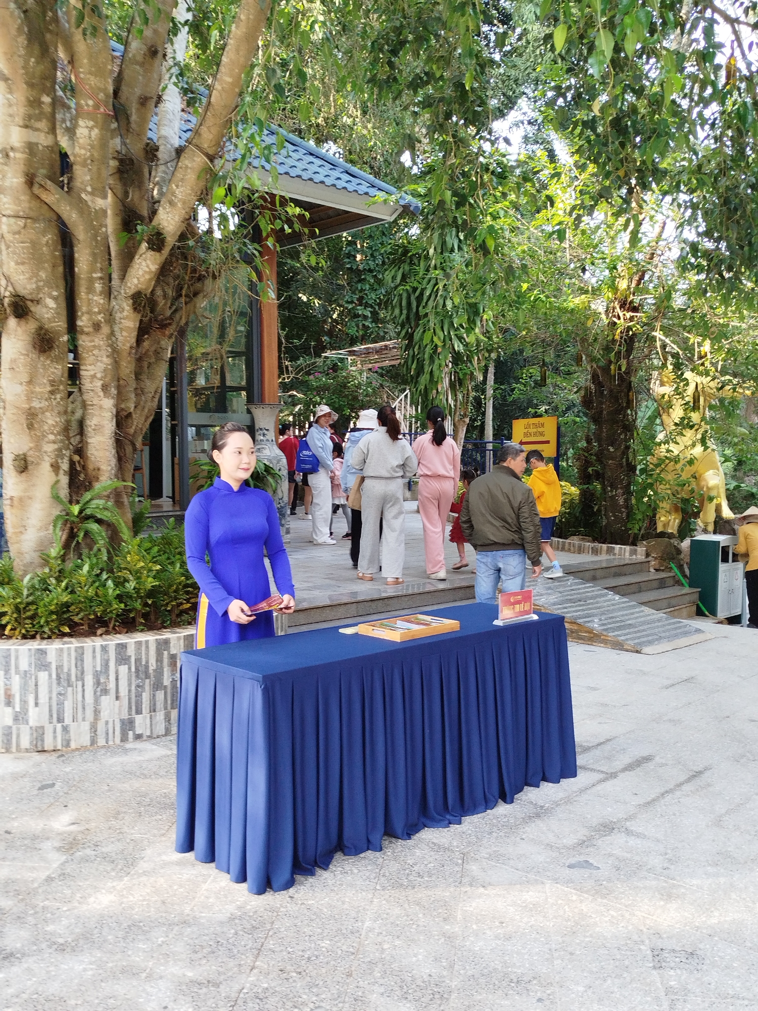 The image captures a scene in Da Lat, Vietnam. A woman in a traditional blue Vietnamese dress (áo dài) stands behind a dark blue draped table. She holds a red and yellow fan. Several people are walking in the background, some heading towards a staircase/ramp that leads to an elevated area marked by a yellow sign. There is a gold statue on the other side of the stairs. The surrounding environment is lush with greenery, including trees and bushes. The architectural style in the background suggests a traditional Vietnamese building. The setting appears to be a tourist attraction or a park.