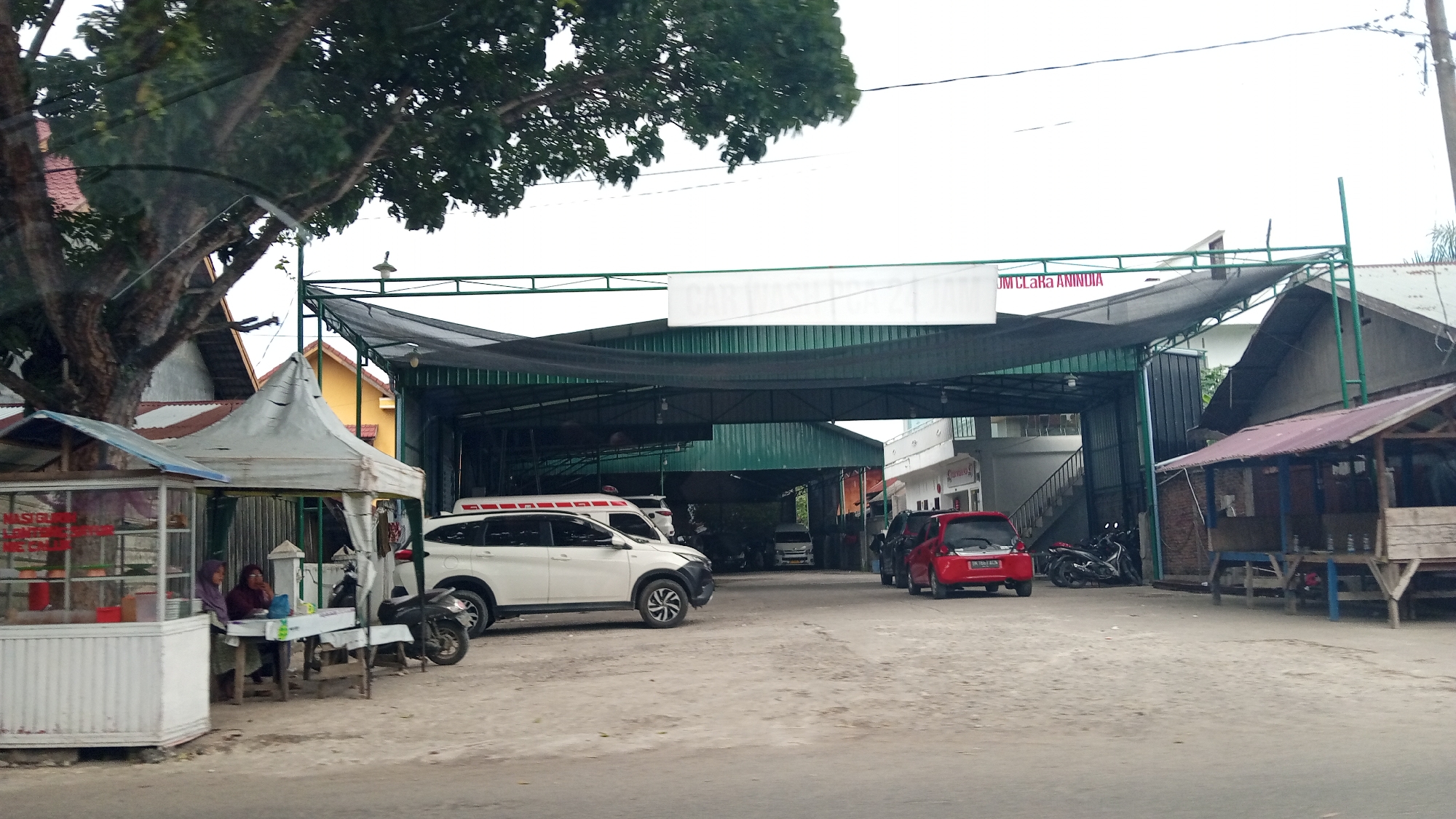 This image captures a street scene in Lhokseumawe, Indonesia, featuring a car wash establishment. In the foreground, a white SUV is parked near a small, open-air food stall. Two women are seated at a table outside the stall, seemingly engaged in conversation or having a meal. A motorcycle is parked behind them. The car wash building is visible in the midground, with a large, dark-colored awning providing cover. Several vehicles, including another white SUV and a red car, are present within or near the car wash area. A sign with text that is partially obscured but appears to include the words "CAR WASH" and "AN INDIA" is attached to the structure above the awning. In the background, a tree with a thick trunk and dark green foliage stands prominently on the left side of the image, partially obscuring some of the buildings. The overall environment suggests a casual commercial area with a focus on vehicle services and local businesses.