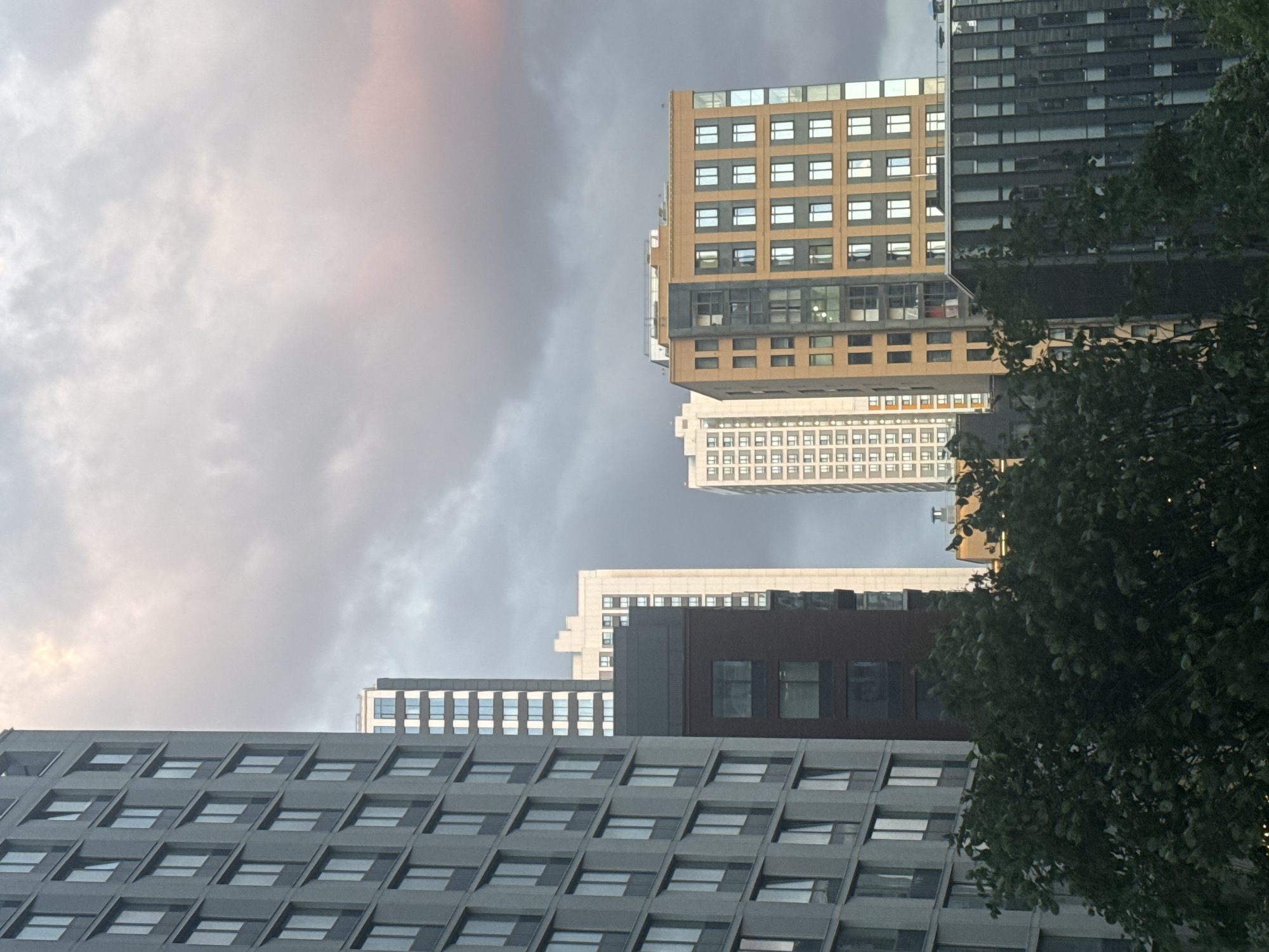 An urban scene featuring a cluster of modern buildings dominates the frame under a cloudy sky. The image appears to be rotated 90 degrees counter-clockwise from a standard upright perspective, showing the buildings stretching horizontally across the view.
In what would be the upper-right section if oriented correctly, a tall building with a light brown or golden-yellow facade features numerous square windows. Below its upper sections, a dark grey, slightly cantilevered section is visible, with more windows. Another building, dark grey or black, with many small, rectangular windows, stands to its right. Further back and slightly below, a white building with an array of small, repetitive windows can be seen.
In the foreground, if viewed upright, a large grey building occupies the lower portion of the frame, characterized by a repeating pattern of recessed, square-like windows. Dark green tree foliage partially obscures some of the buildings on what would be the right side of the frame.
The sky is heavily overcast, with varying shades of grey and white clouds. Hints of a faint pinkish or yellowish light are visible towards what would be the upper-left corner of the sky, suggesting either early morning, late afternoon, or light breaking through the clouds. No people or visible text are present. The scene depicts a modern architectural landscape in Khimki, Russia.