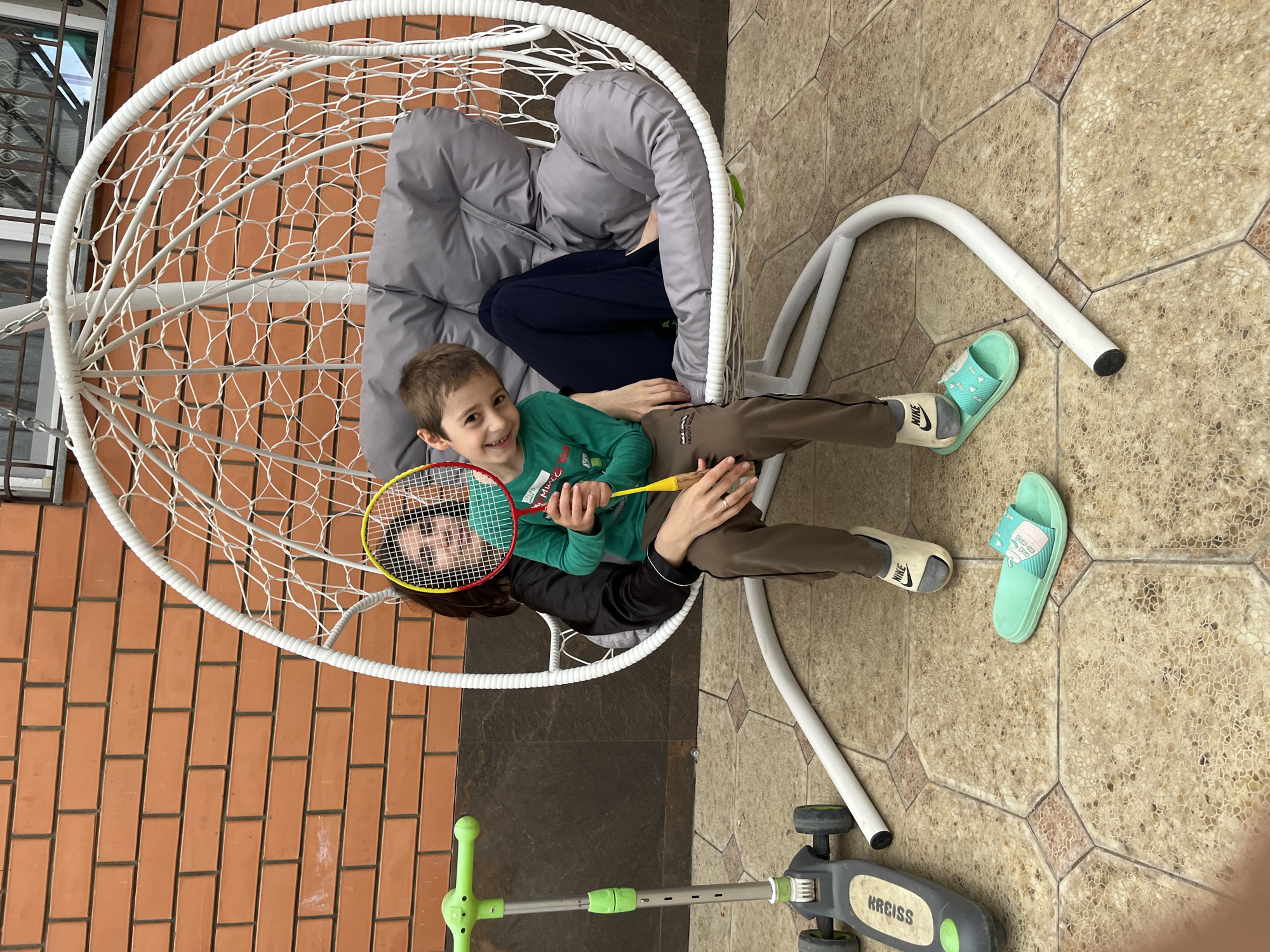 An outdoor scene in Kaspiysk, Russia, depicts a young boy and an adult seated together in a white hanging egg chair. The boy, with short brown hair, wears a long-sleeved green shirt and brown pants, smiling directly at the camera while holding a yellow and red badminton racket. His white Nike slides, worn over grey socks, are visible; one is on his right foot, and the other rests on the tiled ground beside a pair of turquoise slides. The adult, positioned behind the boy, has her face partially visible through the strings of the badminton racket, also smiling. She wears a black long-sleeved top and dark blue pants, with a ring visible on her left ring finger. The egg chair features a grey padded cushion and stands on a white metal frame. To the left, a green and grey three-wheeled scooter, branded "KREISS," rests on the tiled surface. The background consists of a reddish-brown brick wall, with a window featuring a metal security grill visible in the upper right quadrant. The ground is paved with patterned hexagonal and square tiles in shades of beige and terracotta.