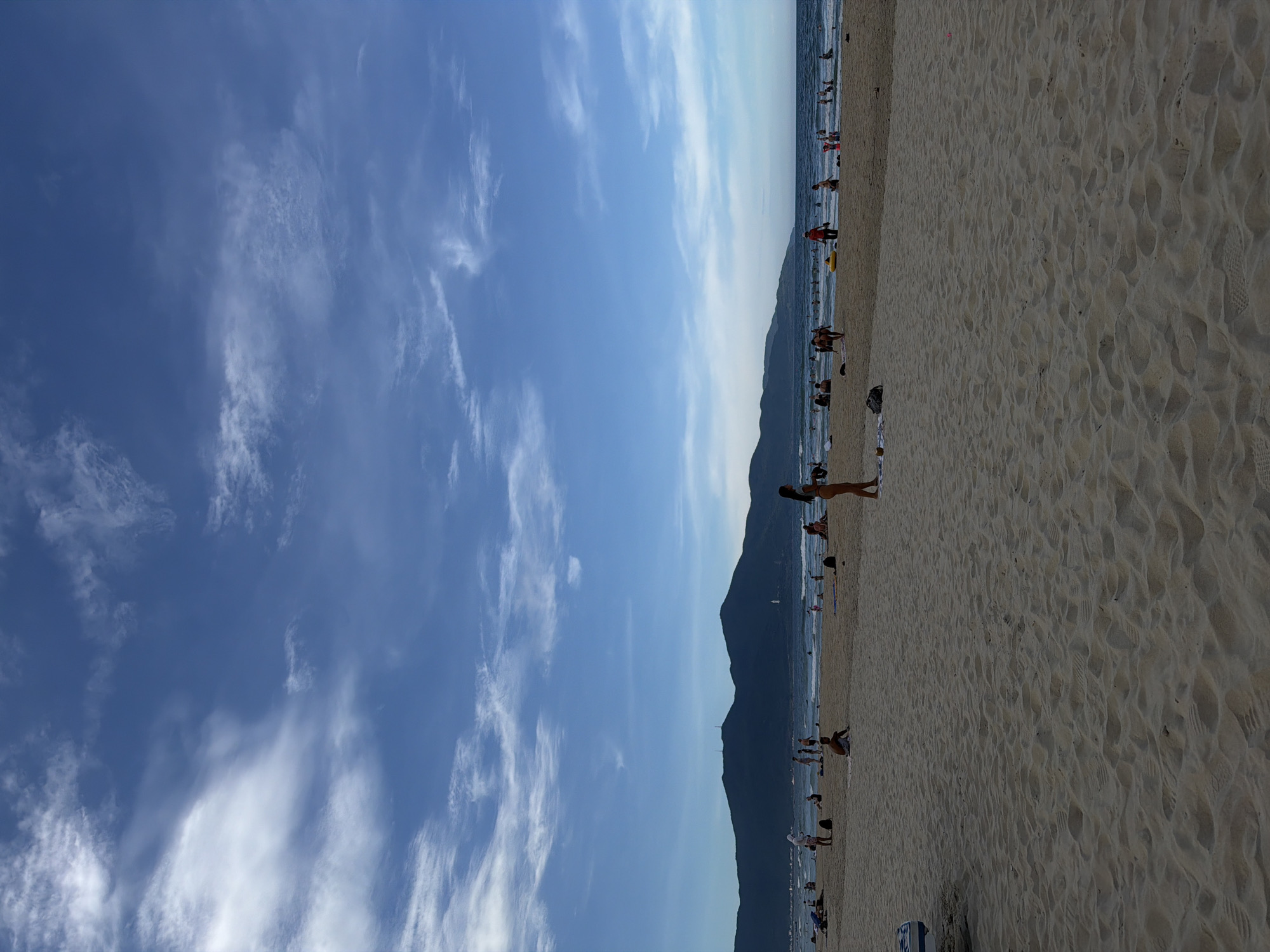 The image shows a beach scene in Đà Nẵng, Vietnam. The sky is predominantly blue with scattered, wispy clouds. A mountain range parallels the coastline. The sandy beach is populated with people, some standing and others sitting or lying down. The beach area is sandy with footprints.