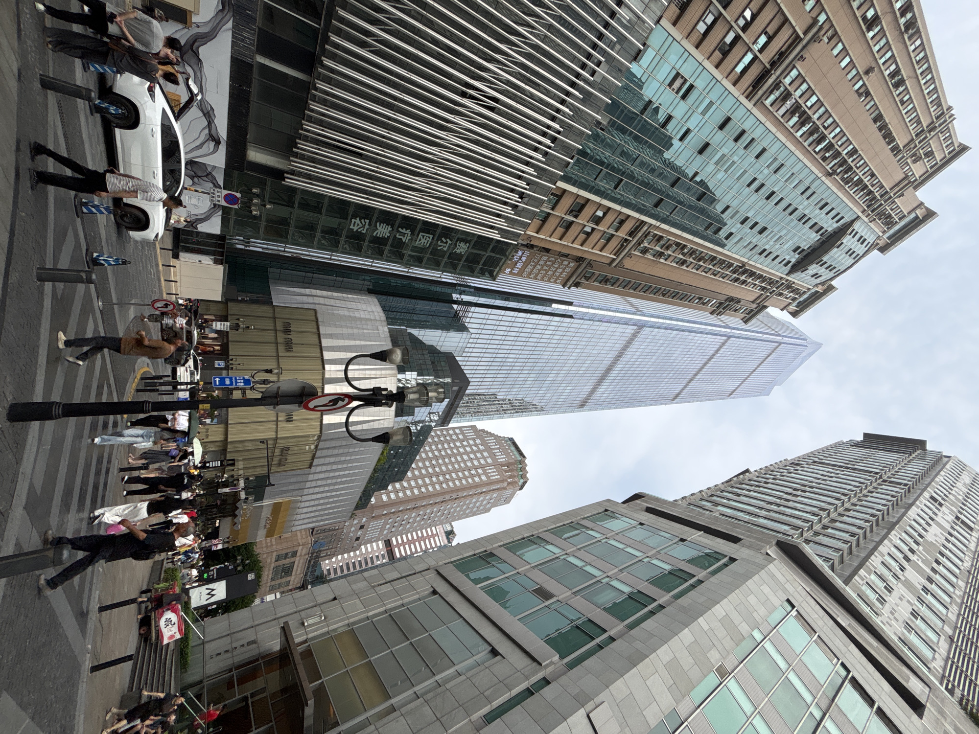 The image captures a street-level perspective in the Yuzhong District of China. The scene is dominated by tall skyscrapers of varying architectural styles, suggesting a dense urban environment. People are walking on the sidewalks and crossing the street, their attire ranging from casual to more formal clothing. A white vehicle is parked near the curb, and a street lamp with multiple signs is visible. The sky is overcast, casting a muted light over the scene. Some signs in Chinese are visible on buildings and street furniture.