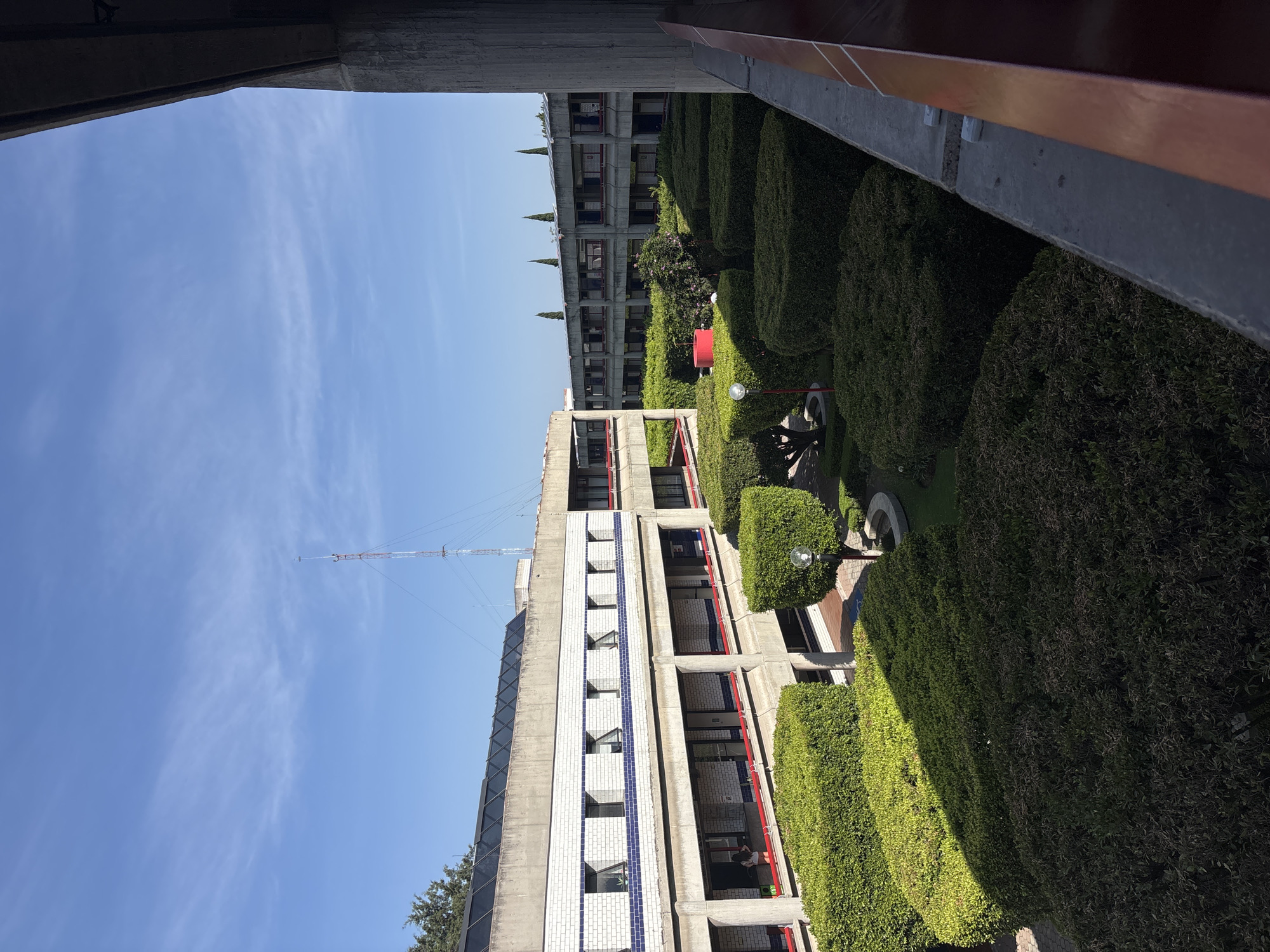 The image depicts a view from an elevated position overlooking a courtyard in San Bernardino Tlaxcalancingo, Mexico. The courtyard contains manicured hedges arranged in a geometric pattern, as well as trees and other greenery. In the foreground, the edges of two building structures are visible, framing the view. One building is constructed with gray concrete, while the other has a red railing. In the center of the frame, a multi-story building is present, featuring a concrete facade with a tiled pattern and red railing on the balconies. A communication tower is also seen extending vertically above the building. The sky is visible above the buildings, exhibiting a clear blue color with scattered white clouds.