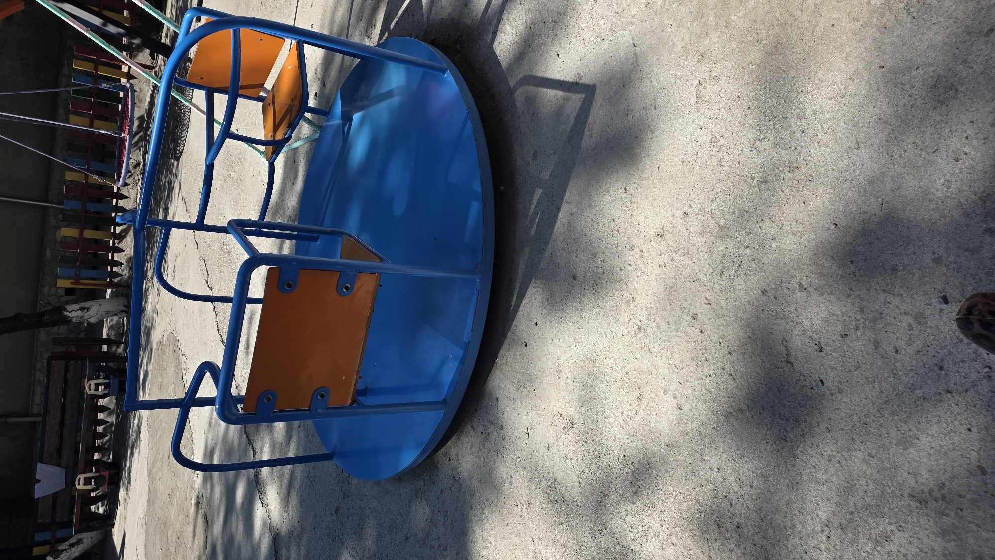 A blue merry-go-round with orange seats is situated on a concrete surface. The merry-go-round is in a playground with a wooden bench and a colorful fence in the background. The ground is dappled with shadows from trees. A portion of a person's foot wearing a brown and black patterned shoe is visible in the bottom right corner of the frame. The scene appears to be in a public park in Tokmok City, Kyrgyzstan.