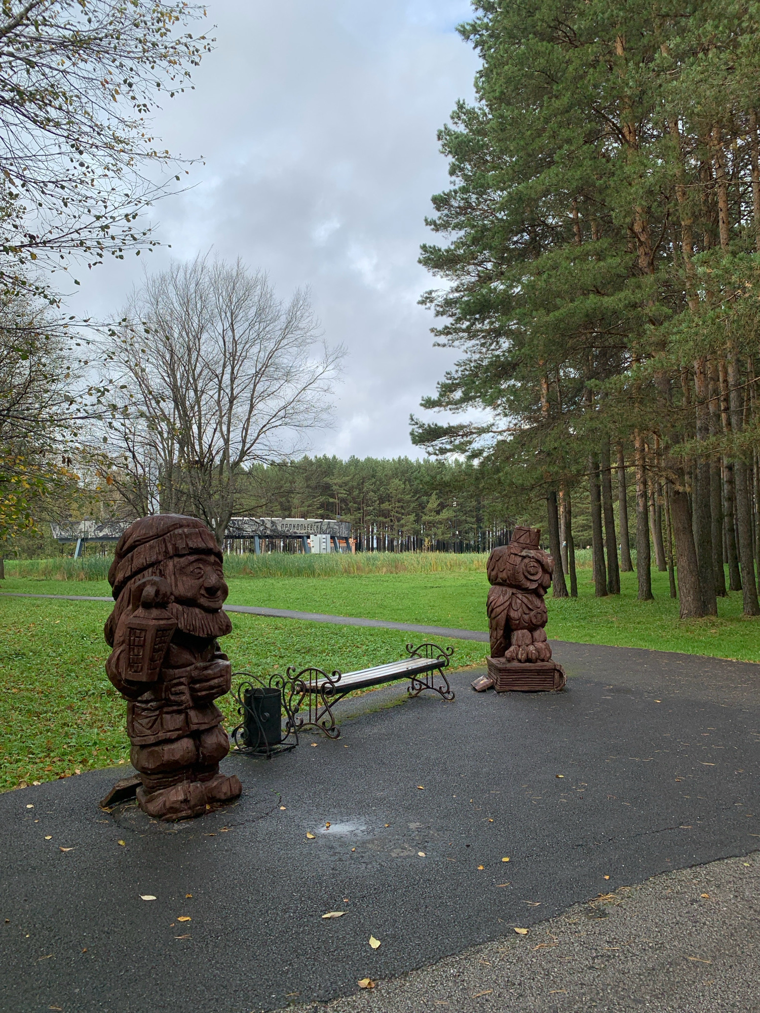 In Prokopyevsk, Russia, a paved path curves through a park on an overcast day. On the left side of the path, a dark brown wooden sculpture of a gnome holding a lantern stands beside a wrought-iron bench and a dark trash receptacle. To the right of the path, another dark brown wooden sculpture depicts a bear-like figure. Beyond the path and sculptures, a wide expanse of green grass slopes towards a line of tall pine trees. In the background, a long, low structure with a bridge-like element spans the distance, with a forest of trees behind it. The overall atmosphere is one of quiet autumn tranquility.