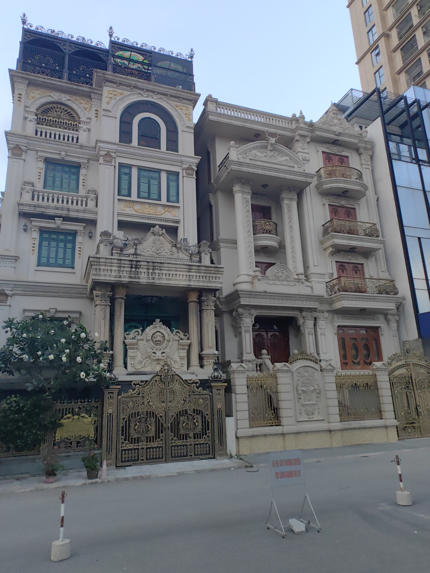 Two ornate, multi-story buildings stand adjacent to each other in Hà Nội, Vietnam. The building on the left is a light color with green-framed windows and decorative gold accents. It has a black iron structure on its top level. The building on the right is also a light color, with multiple balconies and decorative elements featuring a deer. Both buildings have elaborate gold gates. A modern glass building is visible next to the building on the right. The street is paved, and a sign with Vietnamese script stands on a metal easel in the foreground. White posts with red stripes mark the street edge. A small tree is on the left, near the gate of the leftmost building.