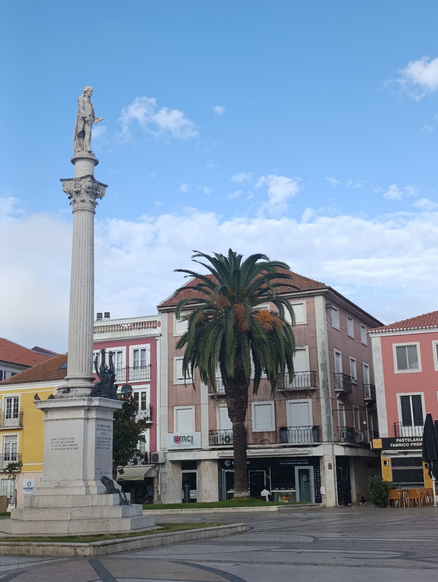 A monument topped with a statue of a man holding a book stands tall in a public square in Setúbal, Portugal. The monument's base is inscribed with text. A large palm tree stands to the right of the monument, with its fronds reaching towards the sky. Behind the monument and palm tree are several buildings, painted in various colors including pink, yellow, and red. The buildings have multiple stories and balconies. The sky is blue with scattered white clouds. In the foreground, the square is paved with patterned tiles. Shops and cafes are visible at the base of the buildings. A few people are present in the square, including one person walking a dog.