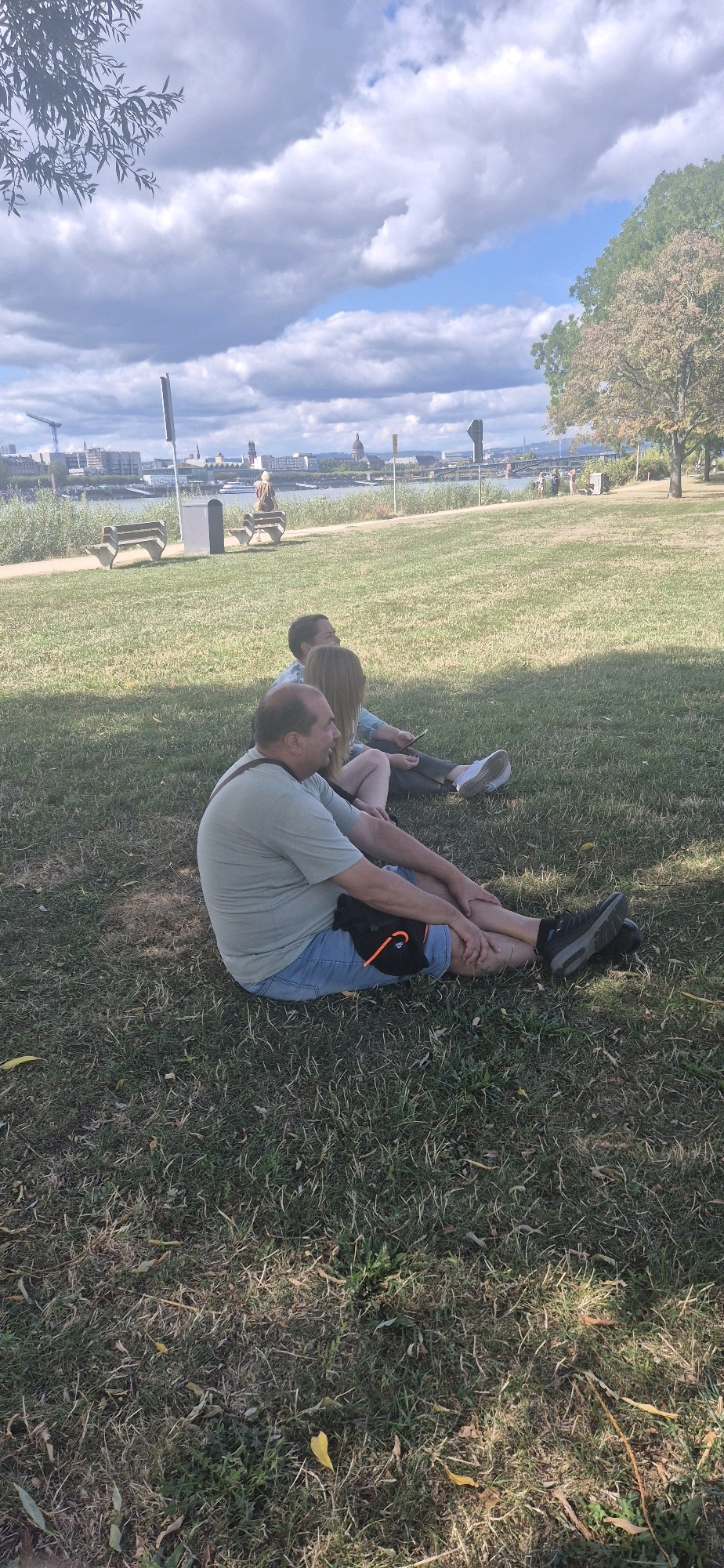 Three individuals are seated on a grassy lawn in a park, facing away from the camera. The person in the foreground, a man wearing a light green t-shirt and blue denim shorts, has a black fanny pack with an orange strap around his waist. He is sitting with his legs extended. Behind him, a woman with long blonde hair is seated next to a man. She is wearing dark pants and a light blue patterned shirt. The man behind her is wearing a light blue patterned shirt. They appear to be looking towards the river and a city skyline in the distance. The city skyline includes buildings, a bridge, and industrial cranes, suggesting an urban setting. The park features scattered trees and benches. The sky is partly cloudy with a clear blue sky visible. The overall scene depicts a relaxed outdoor gathering in a park adjacent to a body of water and a city. This location is likely the Kurpark or a similar park along the Rhine River in Wiesbaden, Germany, given the presence of the river and distinct city skyline with architectural elements.