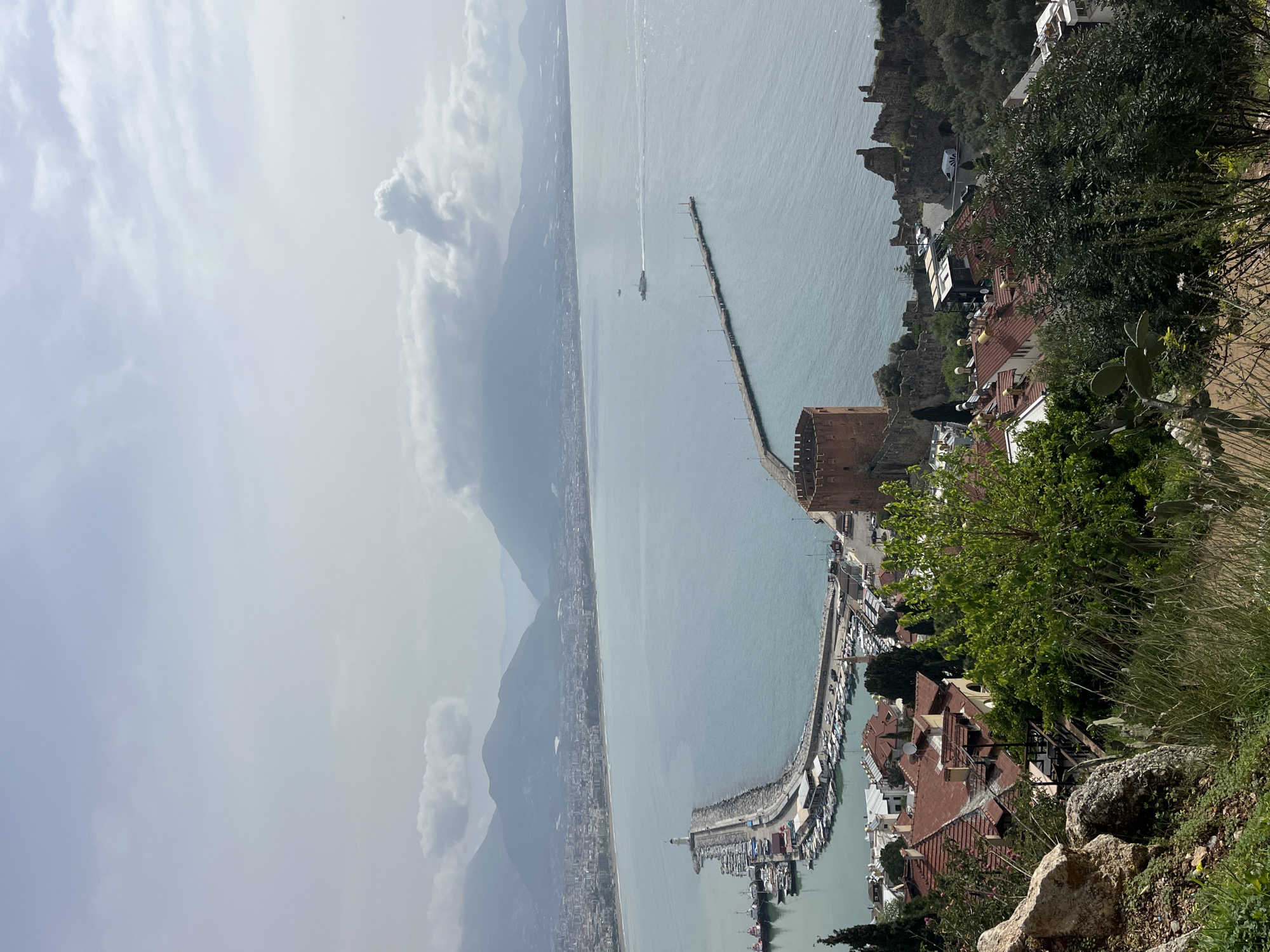 The image presents an elevated, wide-angle view of the Alanya peninsula and its surroundings in Alanya, Turkey, primarily oriented towards the northwest. The sky, occupying the upper third, is overcast with dispersed light grey clouds, allowing some sunlight to reflect on the water.
In the background, a range of hazy, undulating mountains stretches across the horizon, with portions of their upper slopes exhibiting a lighter, possibly snow-covered appearance. Beneath these mountains, the densely built-up urban area of Alanya lines a sweeping coastline with a visible beach.
The midground features the calm, turquoise-blue waters of the Mediterranean Sea. A distinct, curved stone breakwater forms the primary harbor, terminating in a white cylindrical lighthouse. Along the inner perimeter of this breakwater, numerous boats, including smaller pleasure craft and larger, red-hulled tugboats, are moored in a marina. Further into the bay, a long, straight pier with evenly spaced light posts extends from the shore. A white-hulled vessel, possibly a passenger boat, is observed moving away from the harbor, leaving a discernible wake, with another smaller vessel visible further out in the open water. A prominent, octagonal red brick tower, identified as the Kızıl Kule (Red Tower), stands on the coastline near the harbor entrance. Ancient stone walls, likely remnants of the Alanya Castle fortifications, are visible along the rugged eastern shoreline adjacent to the tower.
The foreground consists of a natural, unpaved slope exhibiting rocky outcrops, patches of dry soil, and varied vegetation. This includes light green leafy trees, darker evergreen shrubs, tall grasses, and some succulent plants resembling cacti. Below the immediate viewing point, a cluster of residential or commercial buildings with red-tiled roofs and light-colored facades are nestled among the trees, interspersed with more greenery. No individuals are clearly identifiable or engaged in specific actions within the scene.