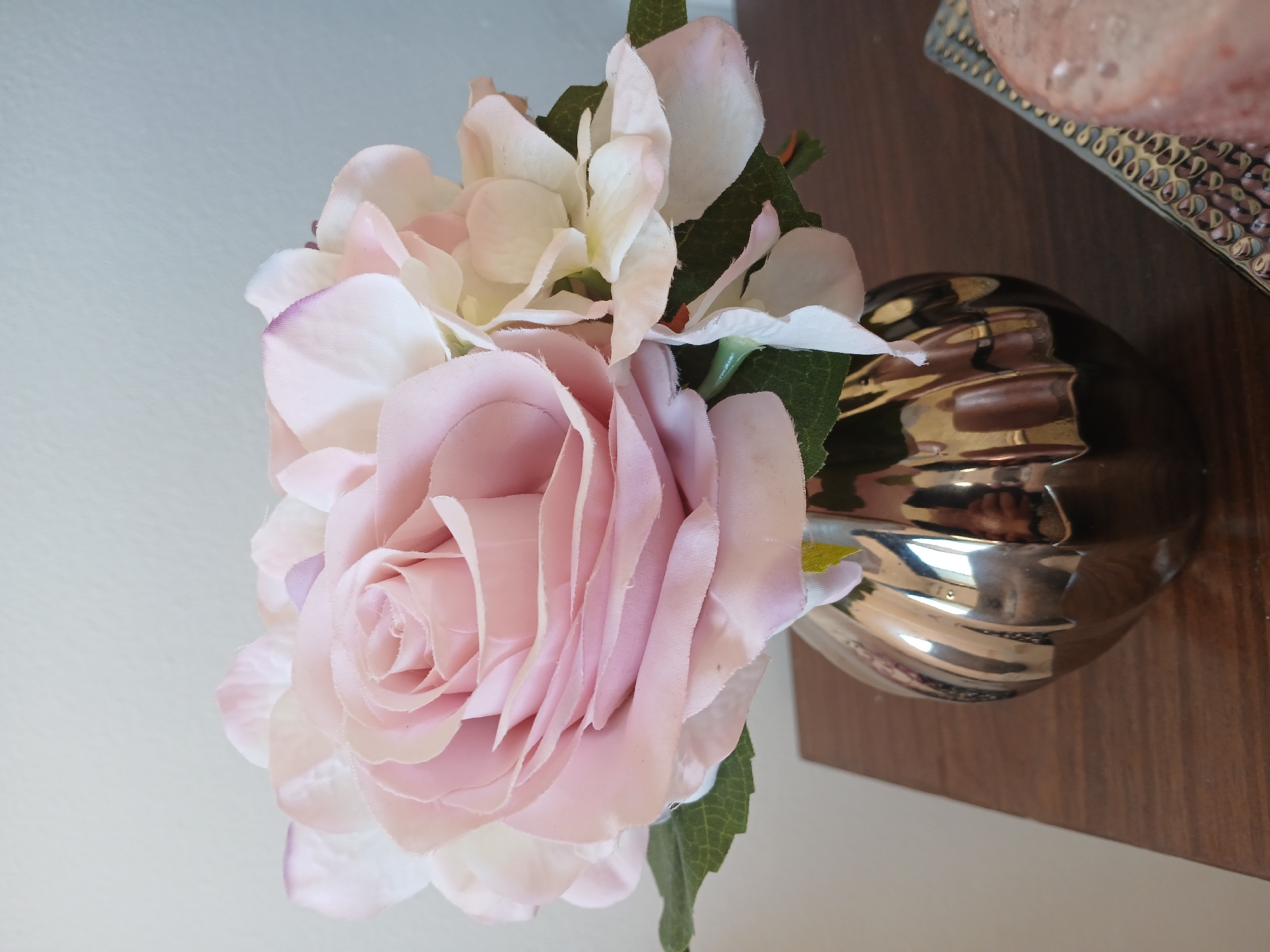A close-up shot displays a bouquet of artificial flowers, primarily pink roses and white hydrangeas, arranged in a reflective, metallic vase. The vase sits on a brown wooden surface, potentially a shelf or table. A portion of a decorative silver tray or dish is visible in the upper right corner of the frame. The background is a plain, off-white wall.