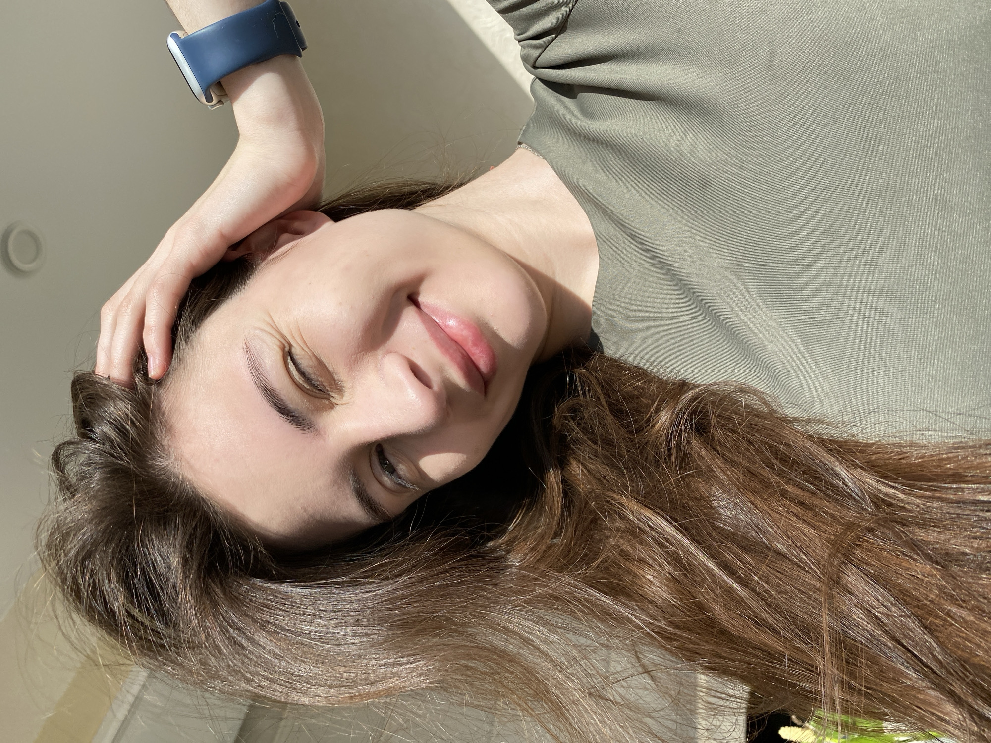 A young woman with long, brown hair is smiling slightly while looking at the camera. She is wearing an olive green top and a blue smartwatch on her left wrist. Her left hand is partially obscuring her forehead. The image appears to be taken indoors in a well-lit environment with sunlight falling on the wall behind her. There is no information available to identify the location as the city of Votkinsk, Russia.