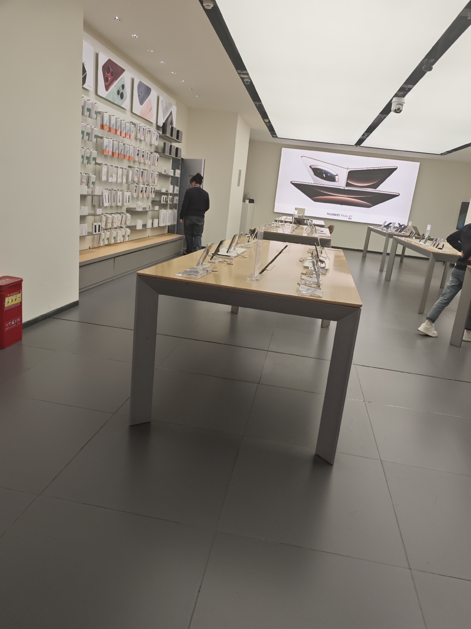 A showroom in Liangjiang, China, features a man in a black jacket examining products on shelves lined with electronic accessories. A table with product displays is in the foreground. Another individual, partially visible, stands at a table further back in the space. A large advertisement display is in the distance. The room has gray tiled floors and is illuminated by overhead lighting.
