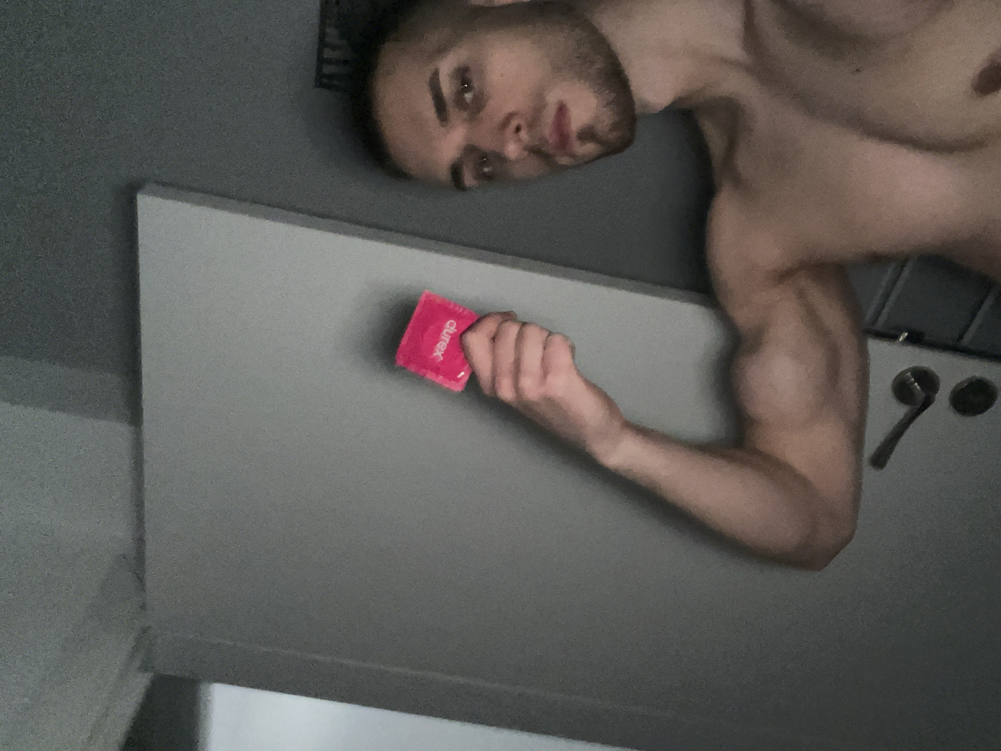 A young adult male, appearing shirtless, is captured in a rotated, overhead shot, looking directly at the camera with a neutral expression. He has short hair and light facial stubble, with his head positioned in the upper left of the frame and his muscular torso and arm extending towards the bottom right. In his right hand, he holds a bright pink rectangular packet with the brand "durex" clearly visible in white text.
The background consists of plain, textured grey surfaces, with a distinct lighter grey, flat surface, possibly a counter or wall, dominating the middle left of the image. Further in the background on the right, two dark circular objects, likely knobs, and a silver, handle-like object are partially visible. The setting appears to be indoors with dim lighting, possibly a bathroom or a minimalist room. There are no direct visual cues within the image to indicate its location in Burgas, Bulgaria, beyond the provided context. The activity is a posed photograph, emphasizing the subject and the product he is holding.