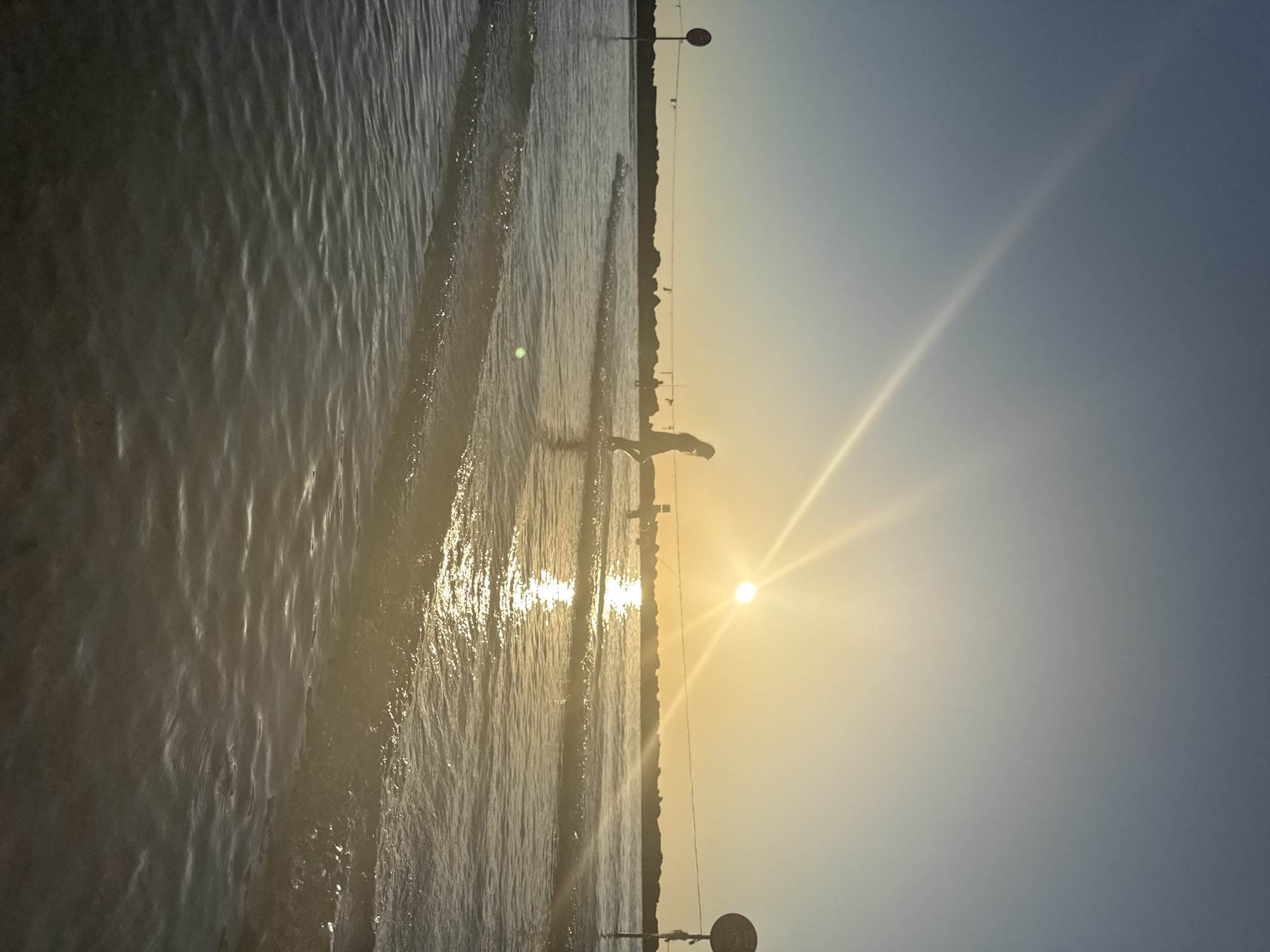 The image shows a serene beach scene at sunset in Netanya, Israel. The sun is low on the horizon, casting a warm golden light across the sky and reflecting on the calm sea. The water is shallow, with gentle waves lapping at the shore. In the foreground, the water is clear, revealing the sandy bottom. A single silhouette of a person is visible in the middle ground, walking near the water's edge. Further in the distance, another person is partially submerged in the water. A rope barrier with buoys marks the swimming area, with a circular sign on a post visible on the left. The background features a rocky breakwater, and beyond that, the tranquil expanse of the sea and sky. The overall impression is one of peacefulness and natural beauty.