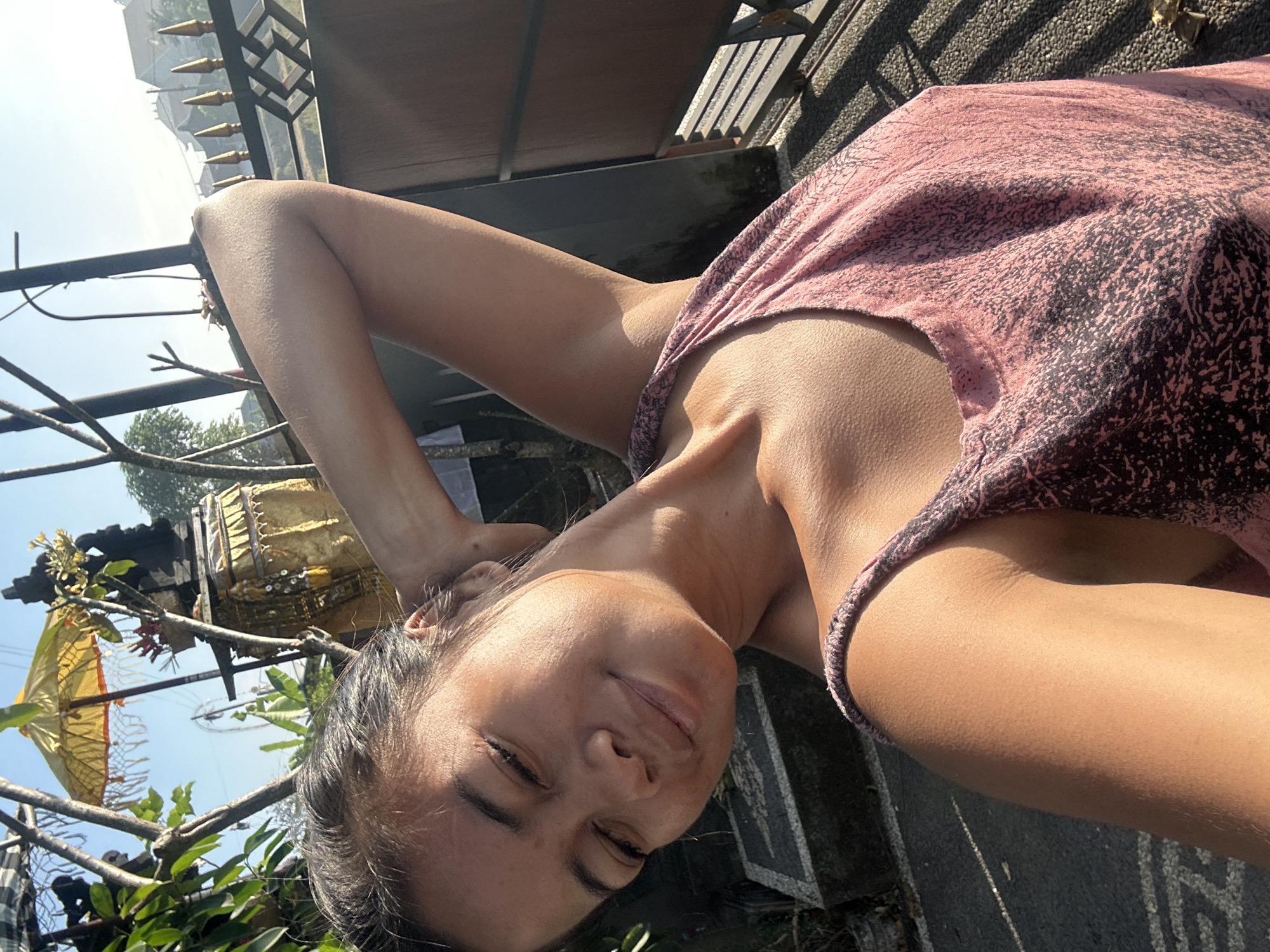A young woman with dark hair pulled back, a slight smile, and eyes closed is captured in a selfie. She is wearing a pink tank top with a black abstract pattern. Her left arm is raised, resting behind her head, showcasing her arm and shoulder. The background suggests an outdoor setting in Sukawati, Indonesia, with lush green foliage, a traditional Balinese umbrella, a shrine structure adorned with gold fabric, and a fence with pointed finials. The lighting indicates it is daytime, with the sun casting shadows.