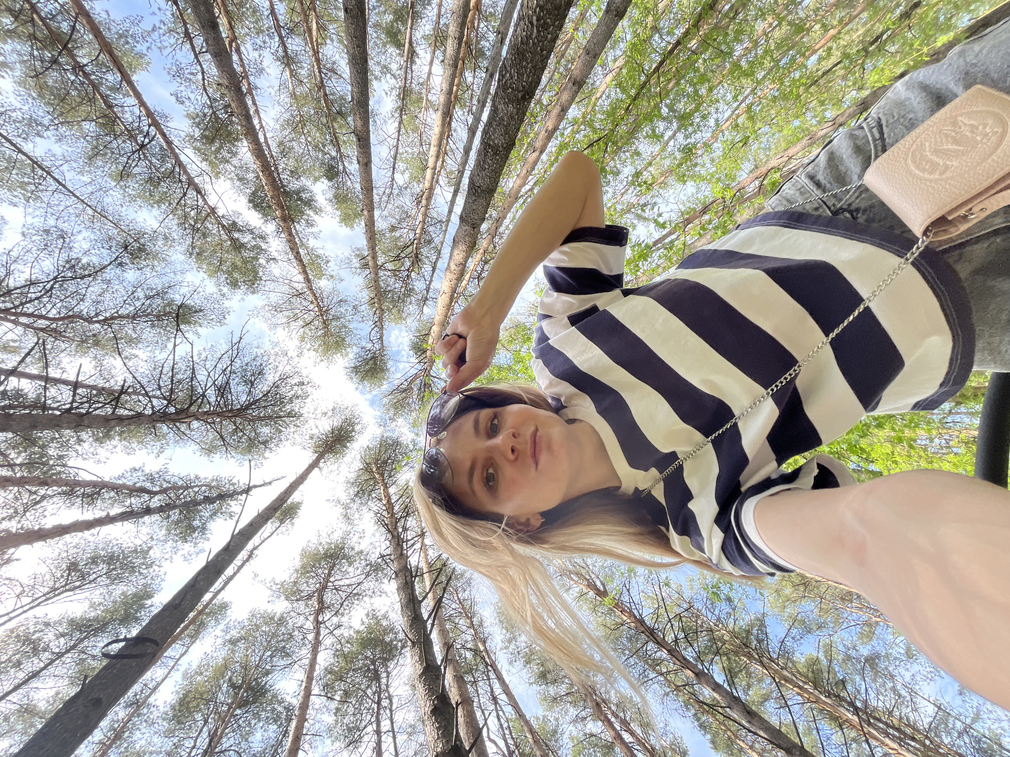 A low-angle, upward-facing shot shows a person, likely female, wearing sunglasses perched on her head, a striped navy and white shirt, and grey jeans, with a light brown small purse visible on the right. The person is positioned against a backdrop of tall, slender trees reaching towards a bright, partly cloudy sky. The trees, with their thin trunks, suggest a forest setting. The photo may have been taken in Chaikovsky, Russia.