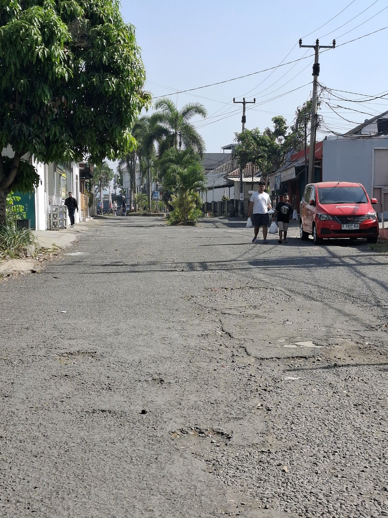 In Karangtengah, Indonesia, a bright, sunny day illuminates a paved road. The road surface is worn and cracked, with visible potholes. On the right side of the road, a bright red car with license plate "D 1017 ALL" is parked. Further down the road, two people, an adult male and a younger male, walk together, both carrying white plastic bags. The adult is wearing a white t-shirt and dark shorts, while the younger male is in a black t-shirt and light-colored shorts.
On the left side of the road, a man in a dark jacket walks away from the camera, approaching a building with white walls. This building appears to be a shop or establishment, with a green door visible. Lush green trees, including a large tree with full foliage and palm trees, line the sides of the road, indicating a tropical or sub-tropical environment. Overhead, electrical wires crisscross the sky, supported by utility poles. The overall scene depicts a casual daytime activity in a residential or commercial street in Indonesia. The clear sky suggests good weather.