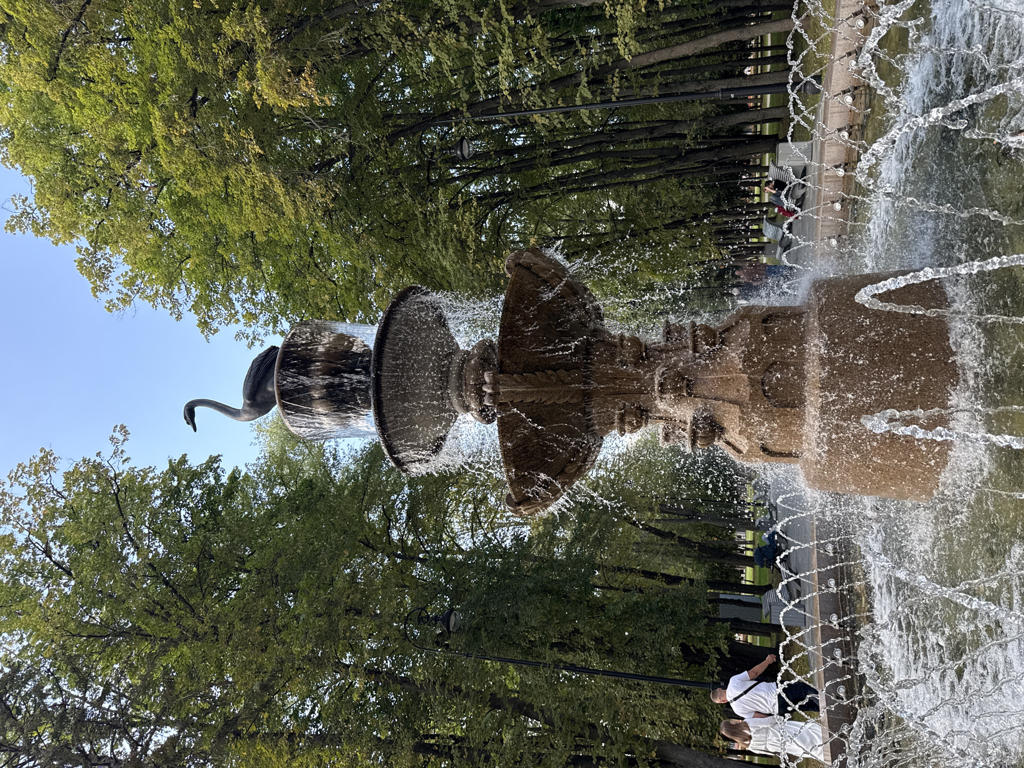 A large, multi-tiered stone fountain with a swan sculpture on top is centrally located in a park, surrounded by lush green trees. Water streams down from each tier, creating a mist. Several people are visible in the background, some walking and others sitting on benches. The scene is lit by natural sunlight, indicating it is daytime. The park appears to be well-maintained, with a paved walkway and lampposts lining the area. This fountain is located in the City of Khimki, Russia.