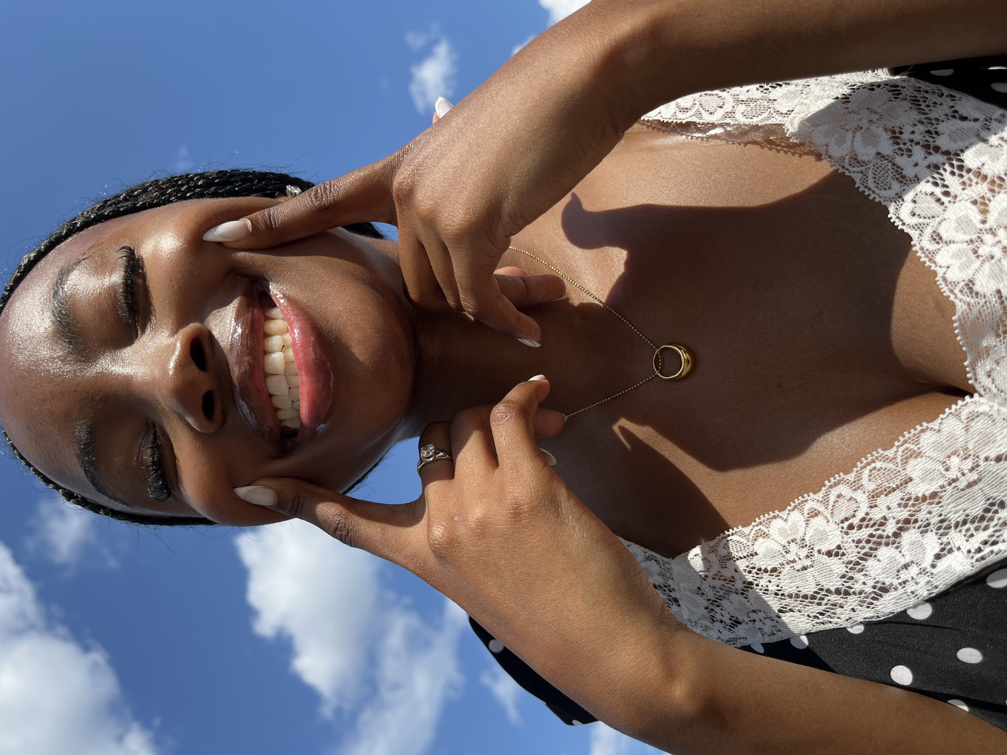A close-up, low-angle shot shows a young Black woman outdoors, smiling widely with her eyes closed and her fingers gently pressing her cheeks. She has dark braided hair, long eyelashes, and is wearing a black and white polka-dot dress with white lace trim. A gold pendant necklace with a circular design rests on her décolletage, and she is wearing a ring on her left ring finger. The background consists of a bright blue sky with scattered white clouds. There are no identifiable landmarks or features that would suggest a specific location within Kazan, Russia, or any other city.